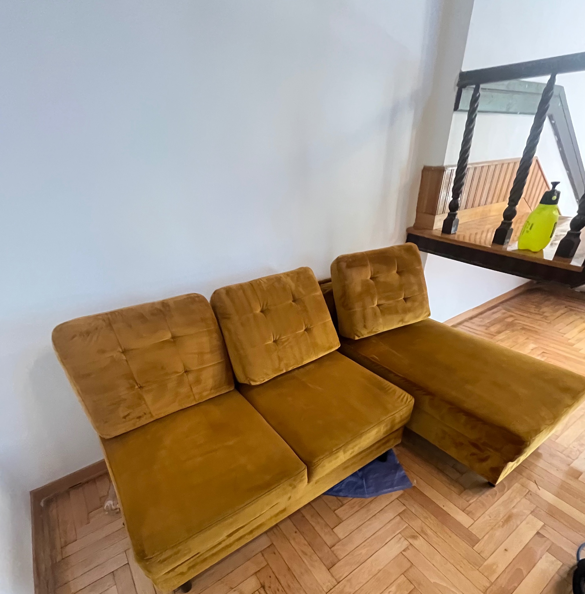 Musztardowa, welurowa sofa modułowa z pikowanymi poduchami, stojąca na parkiecie w jodełkę, obok schodów z drewnianą balustradą i butelką z rozpylaczem.