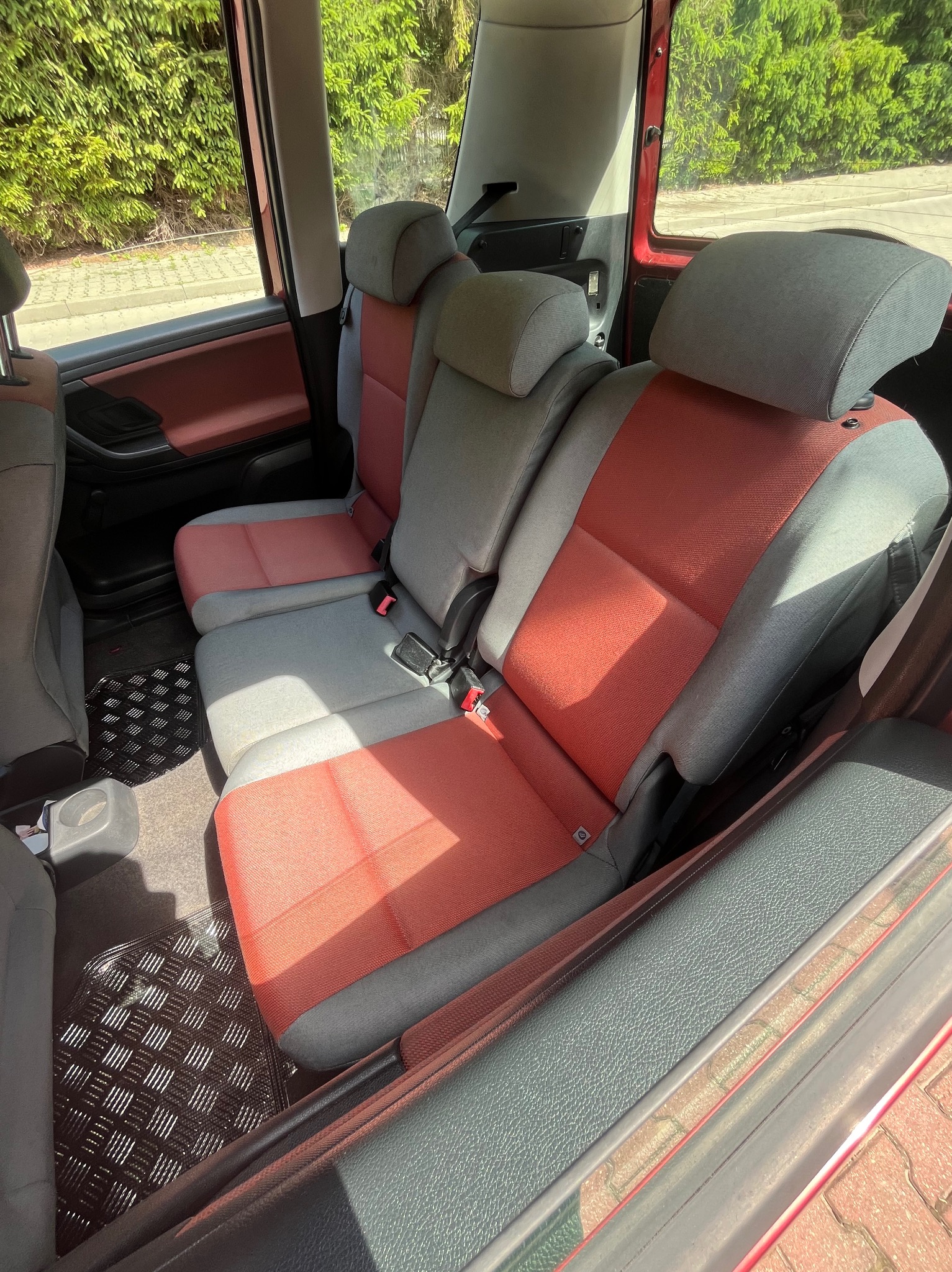 Wnętrze samochodu z trzema rzędami siedzeń w odcieniach szarości i czerwieni, widoczne pasy bezpieczeństwa i gumowa wycieraczka pod nogami pasażerów.