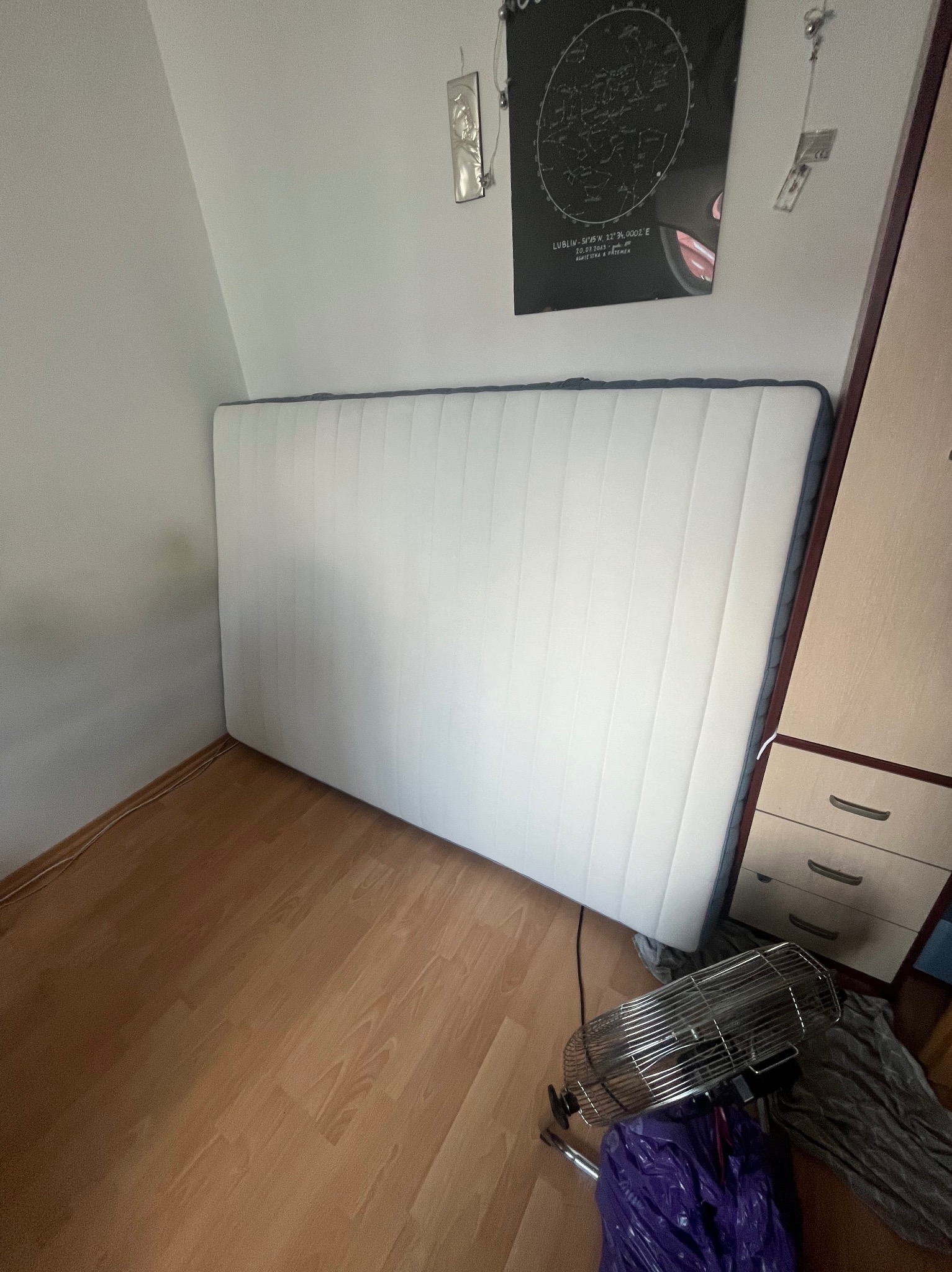 Biały materac piankowy z szarym obszyciem, oparty o ścianę w rogu pokoju z drewnianą podłogą, obok szafki z szufladami i wentylatorem stojącym na podłodze.