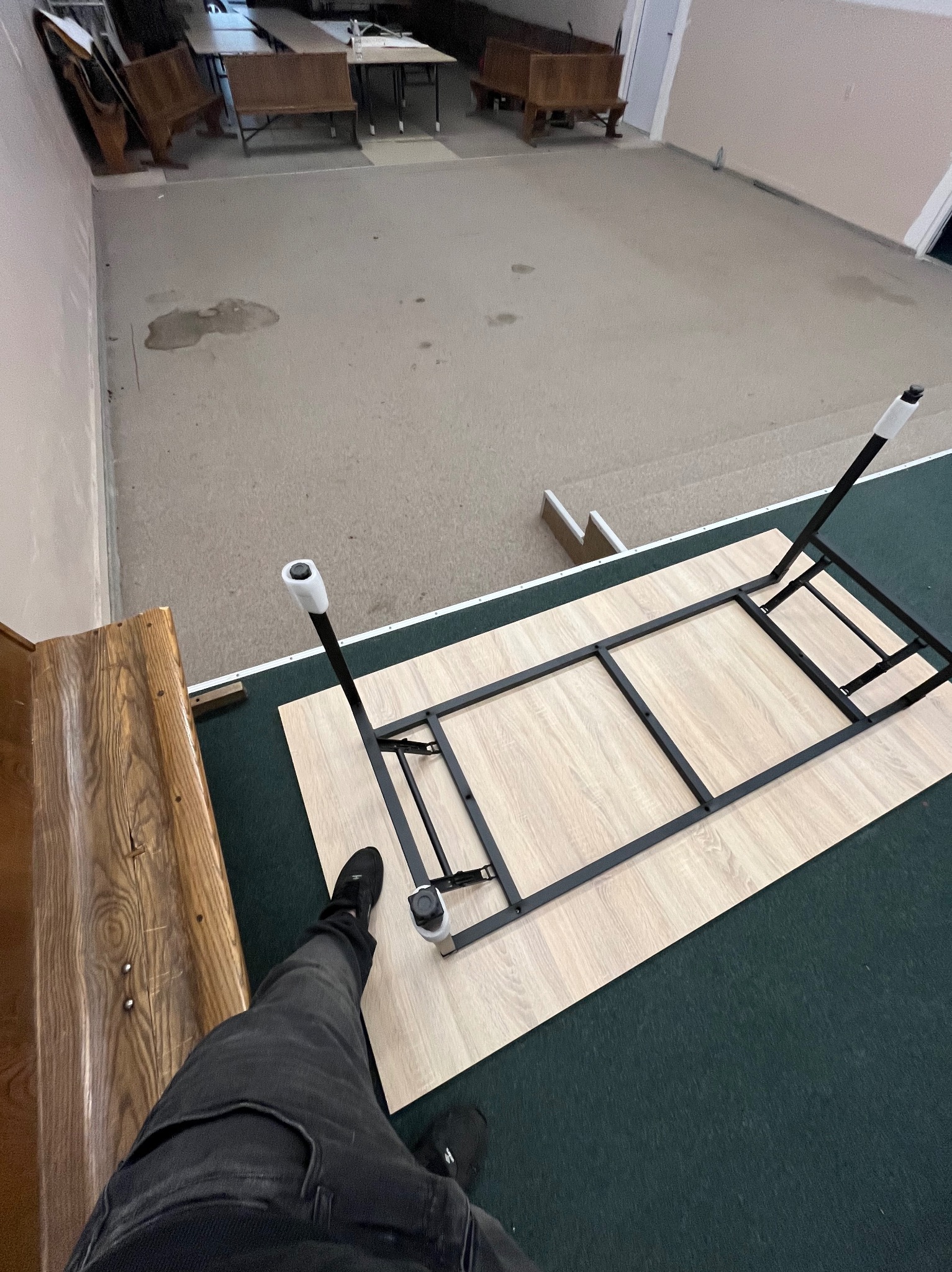 Widok z góry na odwrócony stół z metalową ramą i jasnym blatem, stojący na ciemnozielonym dywanie, obok drewnianej ławki, z widocznymi plamami na podłodze w tle.