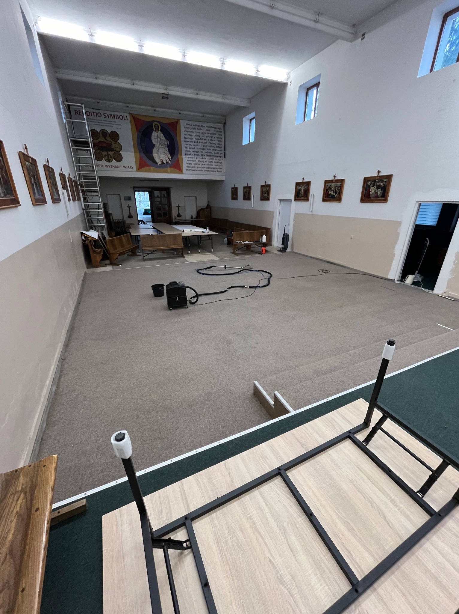 Duża sala z dywanem w trakcie czyszczenia, widoczny odkurzacz piorący i wiązka kabli na dywanie, w tle ściany z obrazami i schody, w rogu sali ustawione stoły i ławki.
