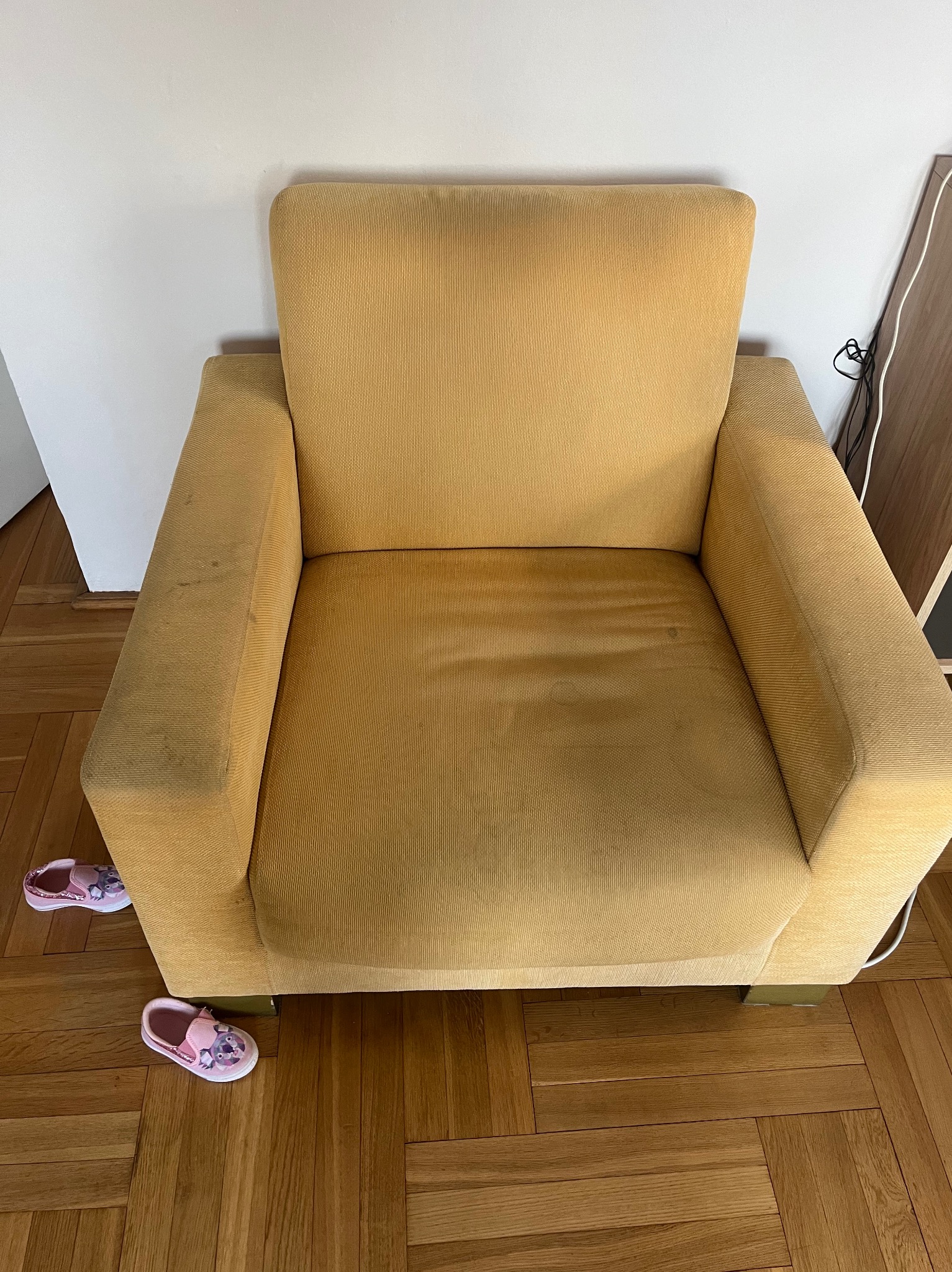 Żółty fotel z widocznymi plamami na podłokietnikach, obok niego para dziecięcych różowych kapci na drewnianej podłodze.