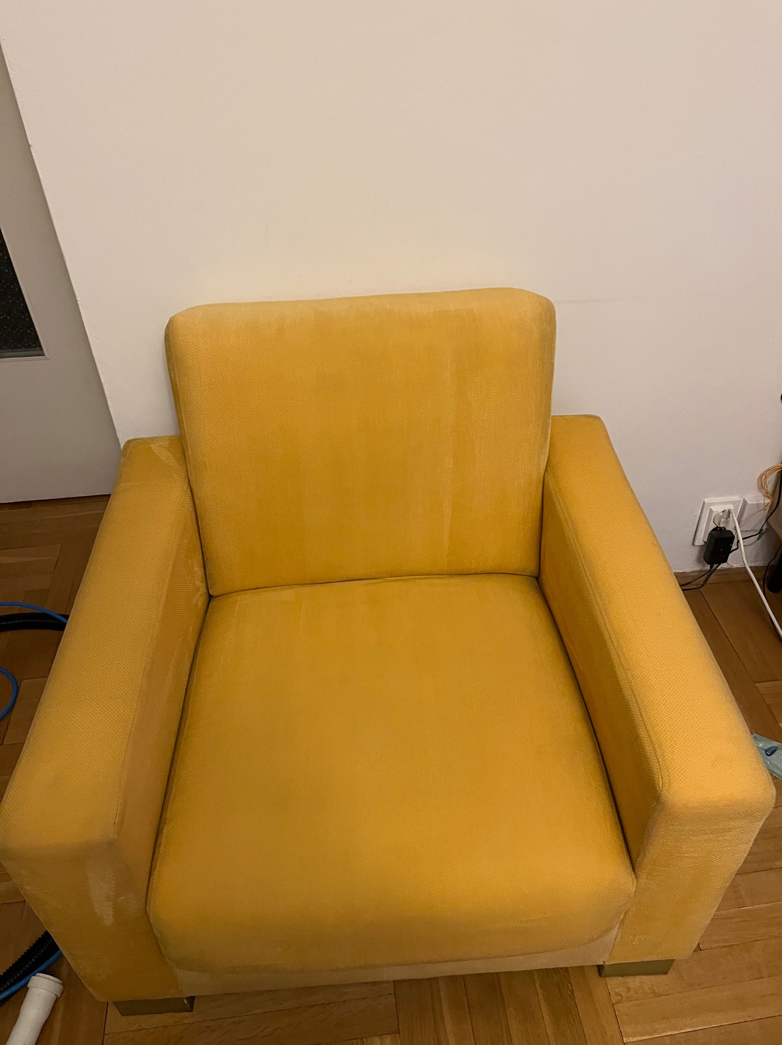 Żółty fotel z plamami na siedzisku i oparciu, stojący na drewnianej podłodze przed białą ścianą, widoczne ślady po czyszczeniu.