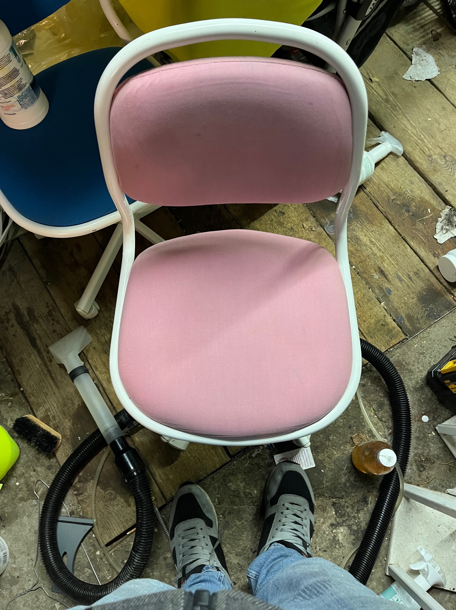 Widok z góry na różowe krzesło z białą ramą, obok sprzęt czyszczący, w tym ssawka odkurzacza i szczotka, na podłodze z desek