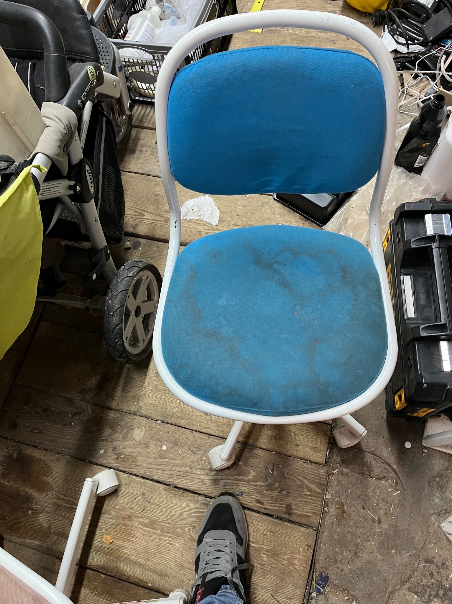Używane krzesło biurowe na kółkach z niebieską tapicerką i białym stelażem, widoczne plamy na siedzisku, otoczone różnymi przedmiotami w zagraconym pomieszczeniu, fragment buta osoby fotografującej...