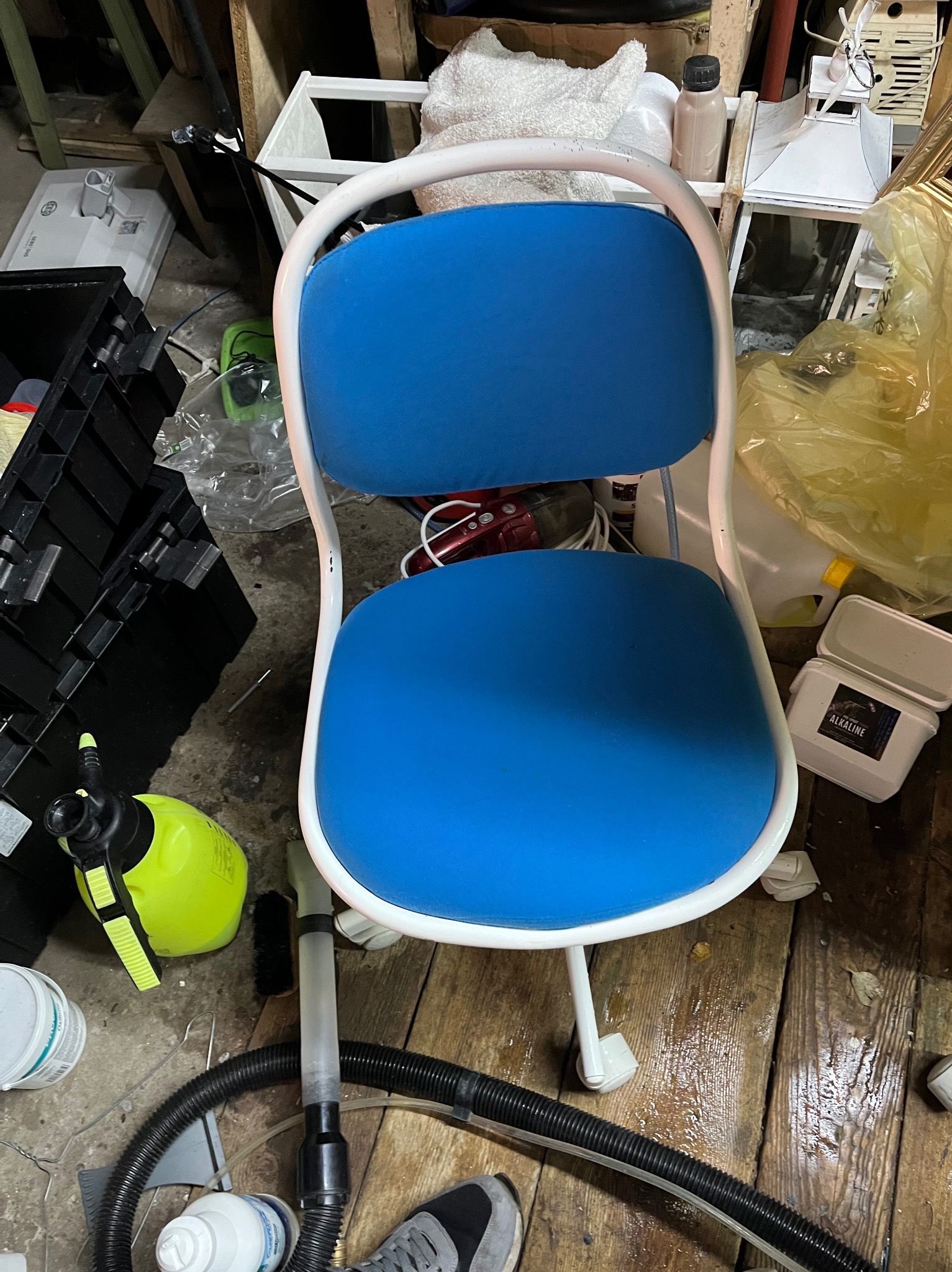 Niebieskie krzesło biurowe na kółkach, otoczone sprzętem do czyszczenia, w tym żółtym spryskiwaczem i wężem odkurzacza, na tle drewnianej podłogi i zagraconego otoczenia.