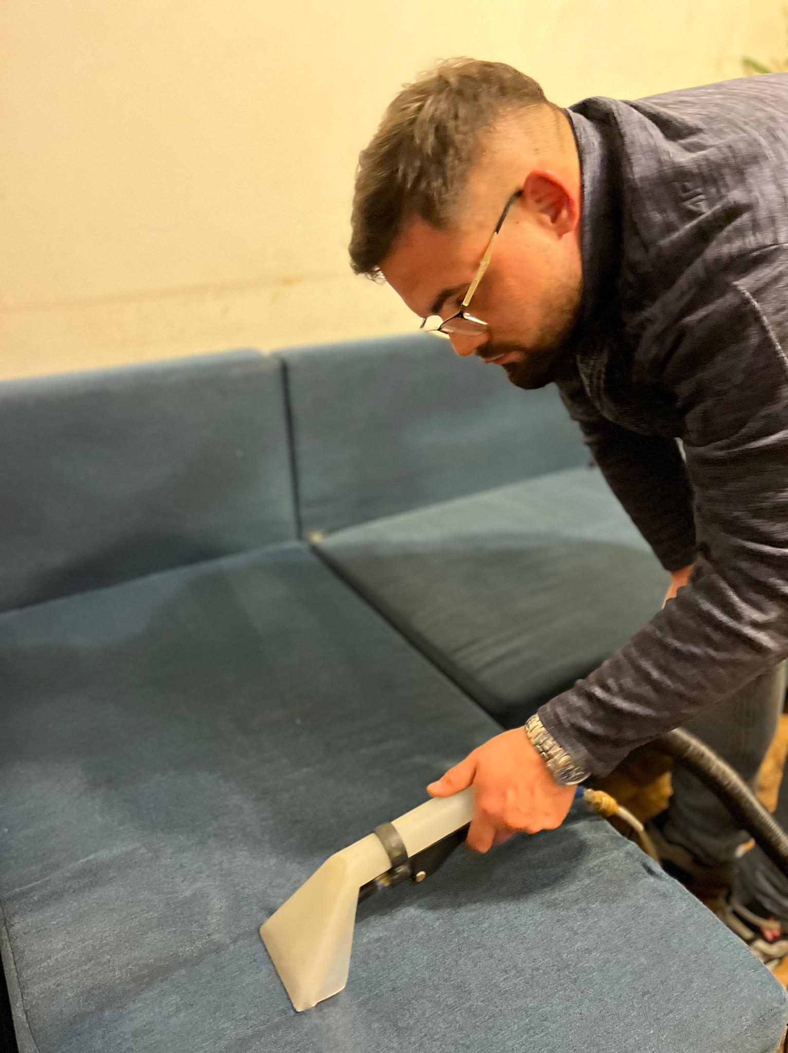 Mężczyzna w okularach czyści niebieską kanapę za pomocą profesjonalnego odkurzacza z przezroczystą końcówką.