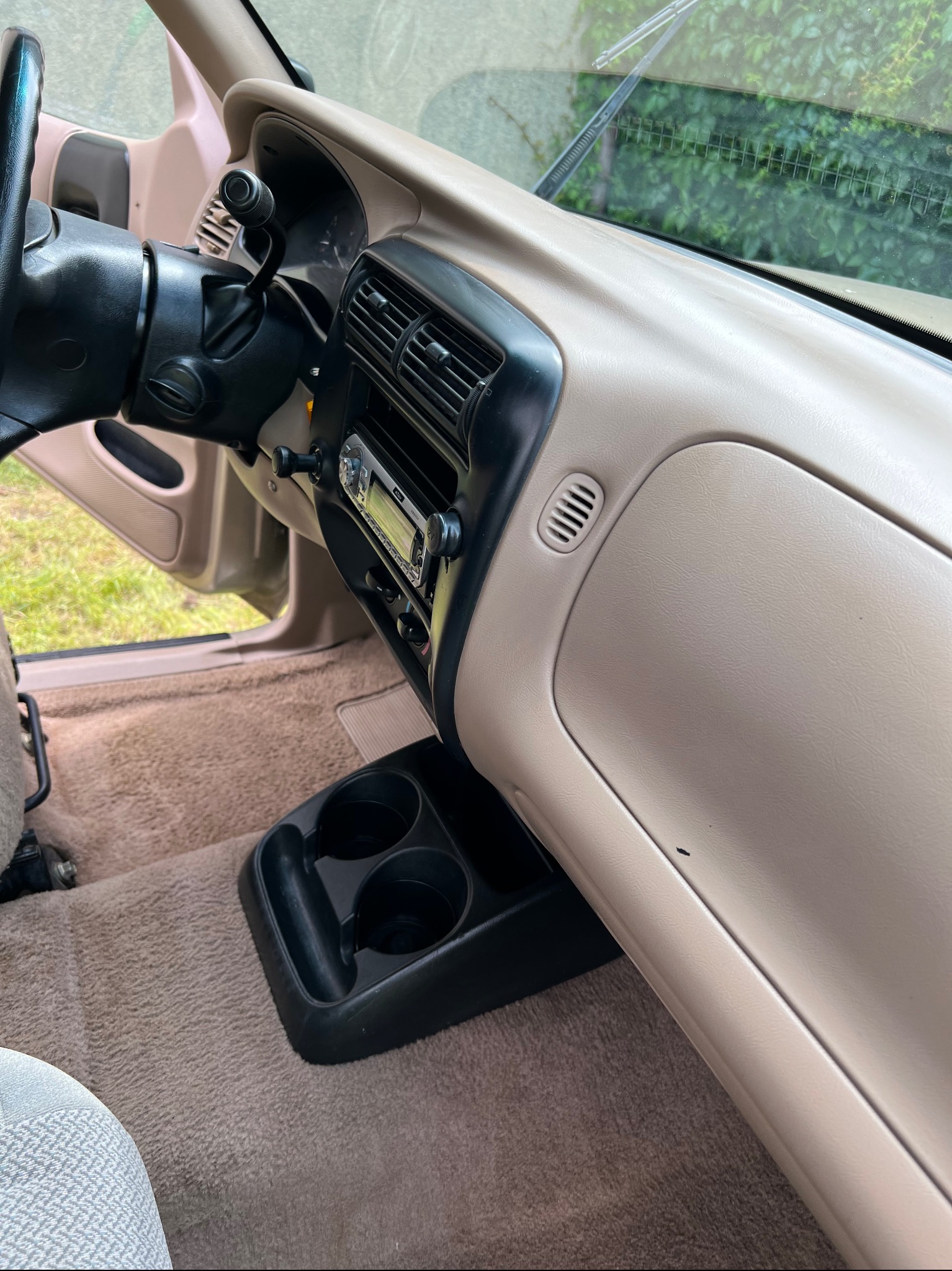 Wnętrze samochodu z beżową deską rozdzielczą, widoczna kierownica, radio samochodowe, kratki wentylacyjne i uchwyt na napoje, z zewnątrz widoczna zieleń.