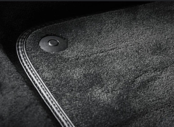 Szary dywanik samochodowy z ciemnoszarym mocowaniem na podłodze i obszyciem z czarno-białą nitką.