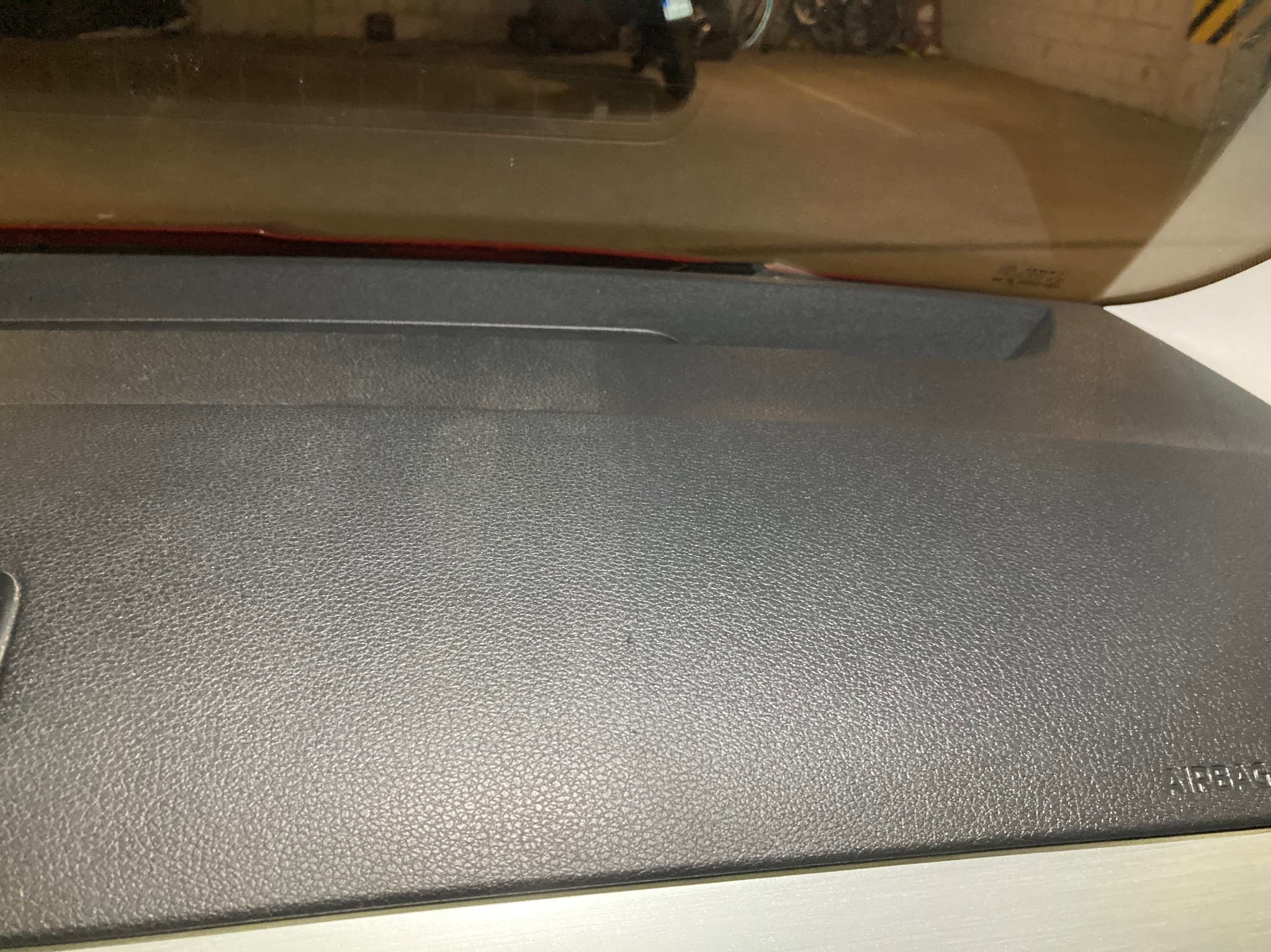 Szara, teksturowana deska rozdzielcza samochodu z widocznym napisem 'AIRBAG' w prawym dolnym rogu, odbicie parkingu w przedniej szybie.