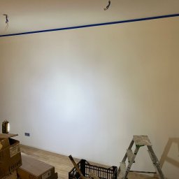 Karol Bal - Przygotowana do malowania ściana w pomieszczeniu, zabezpieczona niebieską taśmą malarską wzdłuż sufitu i narożników, widoczne kartony z materiałami budowlanymi, drabina i oświetlenie warsztatowe