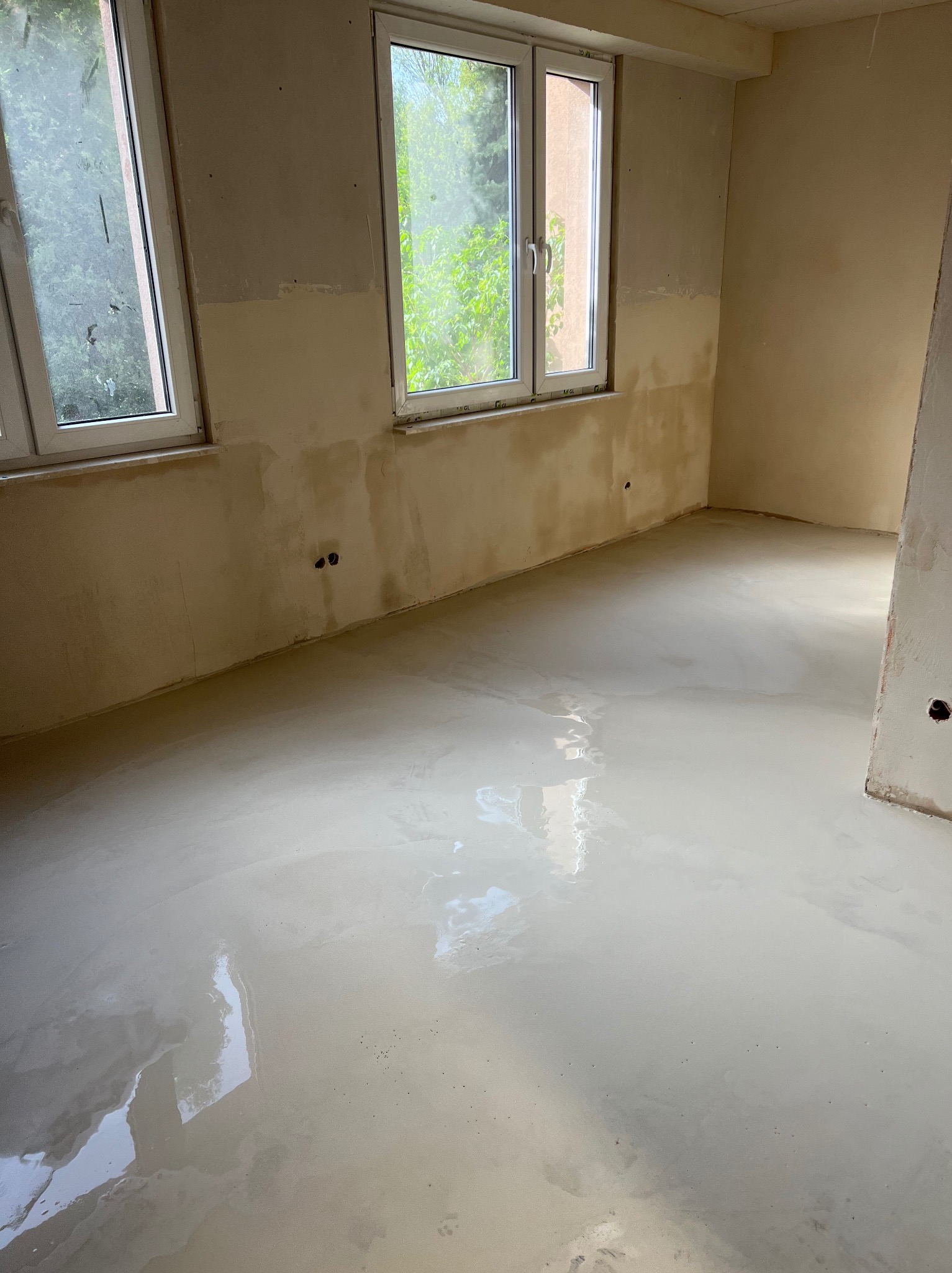 Pokój w trakcie prac wykończeniowych: świeżo wylana, błyszcząca posadzka samopoziomująca, ściany częściowo przygotowane pod malowanie, widoczne okna z widokiem na zieleń.