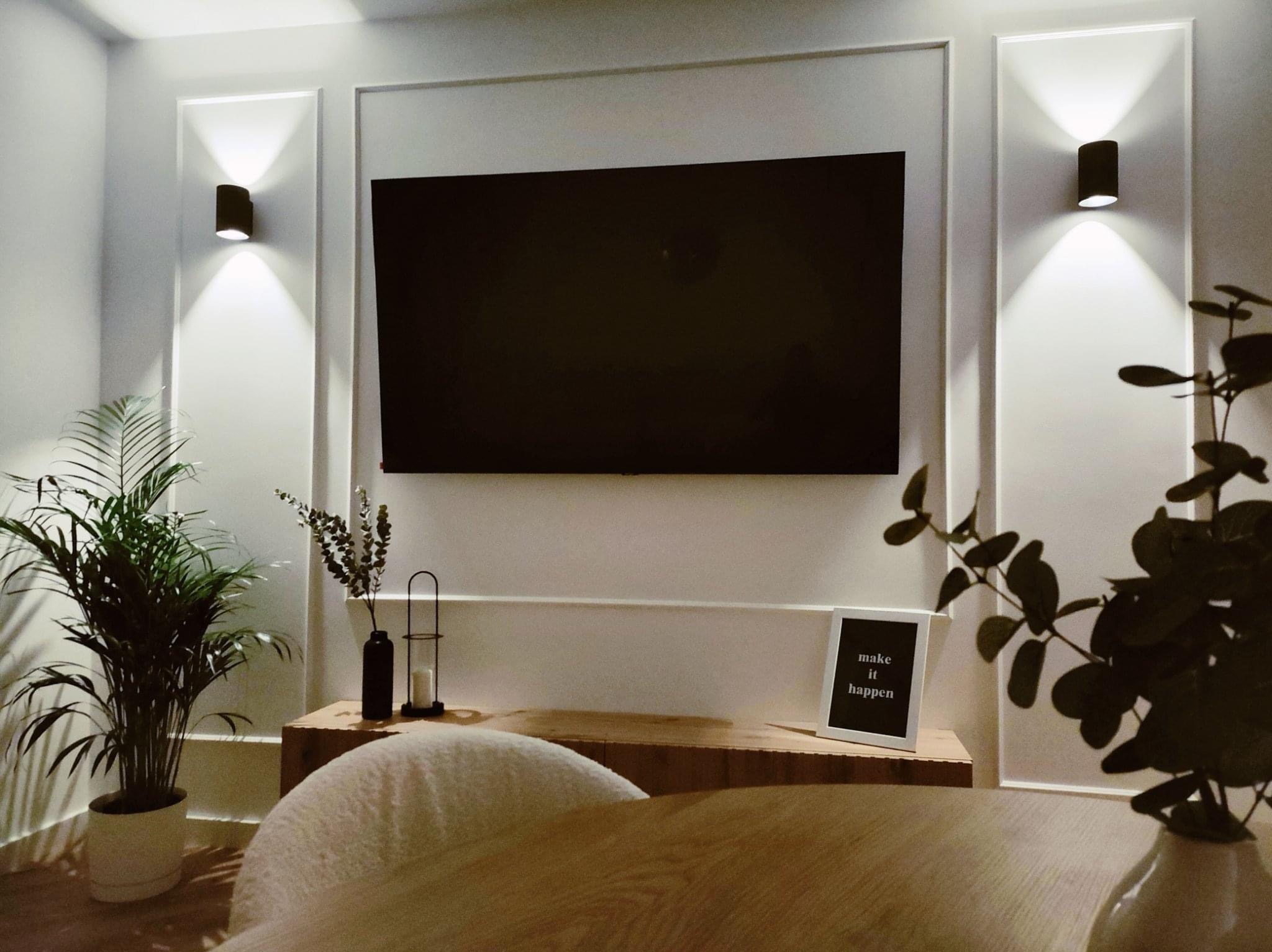 Nowoczesne wnętrze salonu z czarnym telewizorem na ścianie z dekoracyjnymi listwami, oświetlone kinkietami, z roślinami doniczkowymi i drewnianą komodą z dekoracjami.