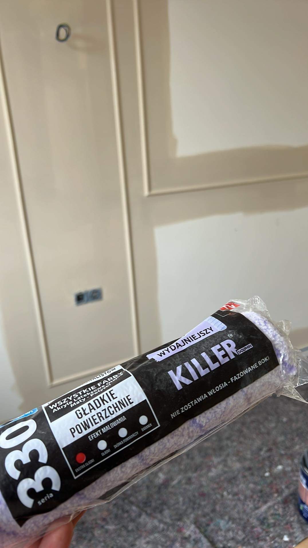 Wałek malarski 'Killer' w folii na tle świeżo pomalowanej kremowej ściany z dekoracyjnymi listwami i gniazdkiem elektrycznym.