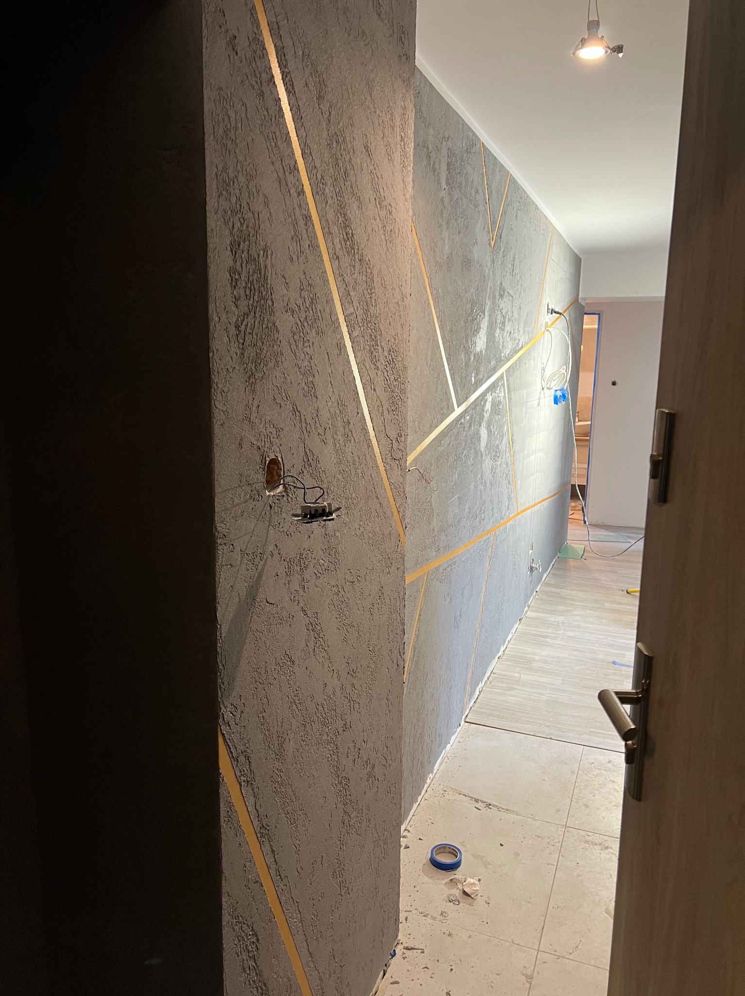 Korytarz w trakcie prac wykończeniowych: ściany pokryte szarą fakturą z geometrycznymi złotymi paskami, widoczne niedokończone gniazdko elektryczne, zabezpieczona podłoga i otwarte drzwi.