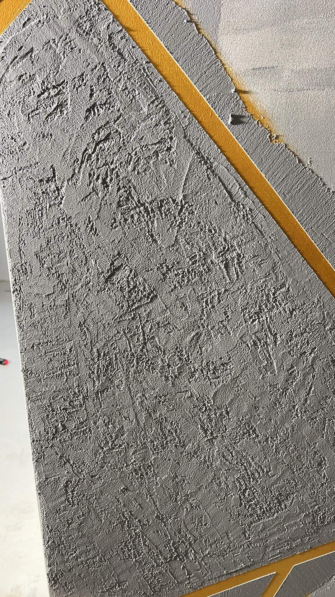 Szara ściana z wyraźną fakturą i geometrycznym wzorem z cienkich, złotych pasków.