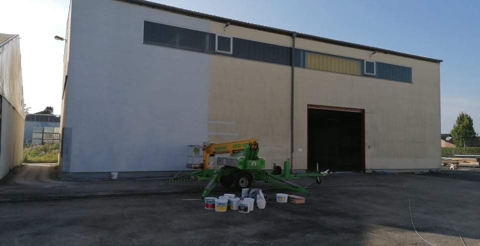 Częściowo pomalowana ściana zewnętrzna budynku przemysłowego z podnośnikiem koszowym i puszkami farb na ziemi.