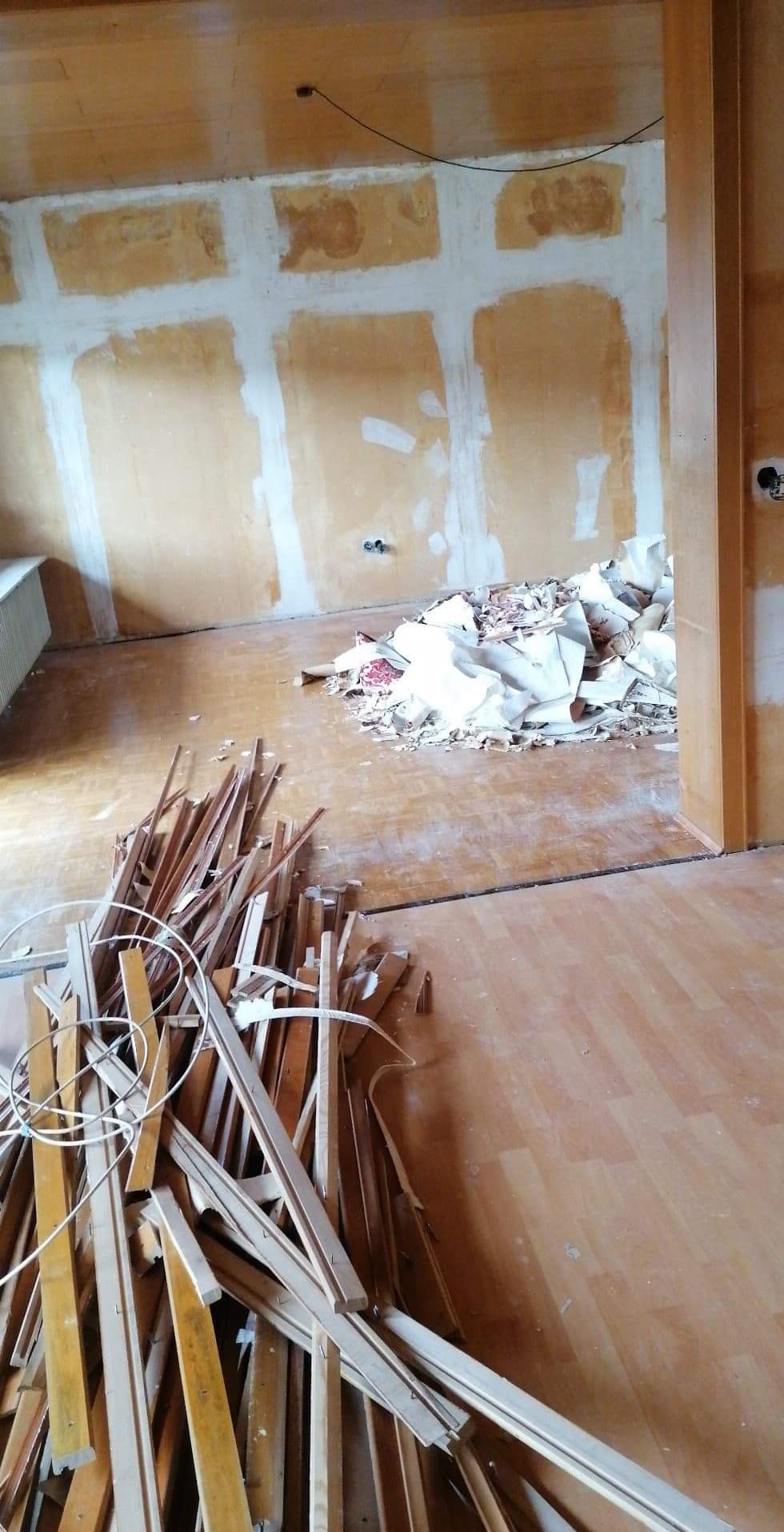 Pokój w trakcie remontu: fragmenty usuniętej tapety, ślady po szpachlowaniu na ścianach, sterta drewnianych listew przypodłogowych na podłodze.