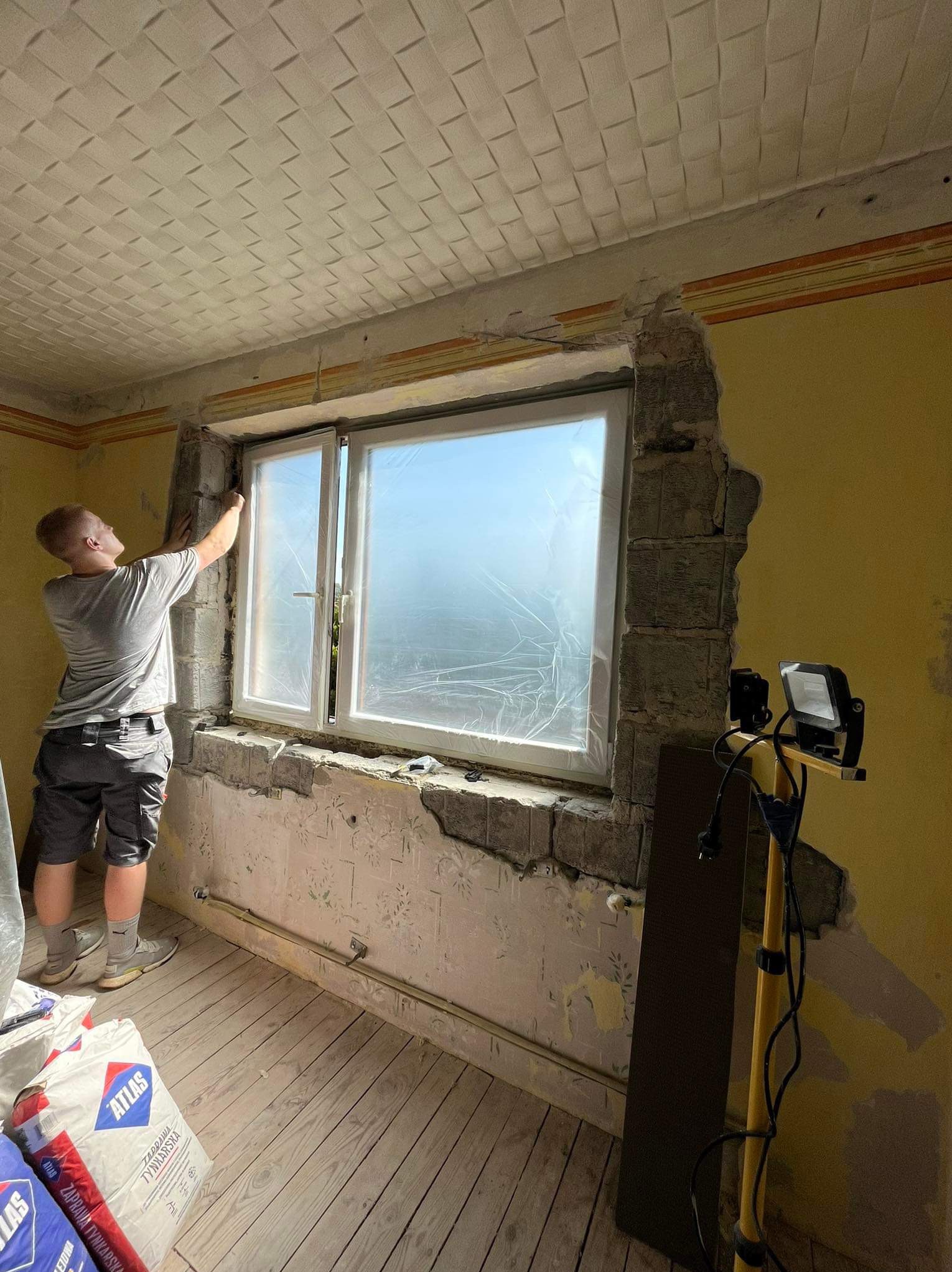 Pracownik przygotowuje ościeże okienne do prac wykończeniowych, widoczny fragment ściany z odsłoniętym murem, worki z zaprawą tynkarską Atlas oraz lampa budowlana.
