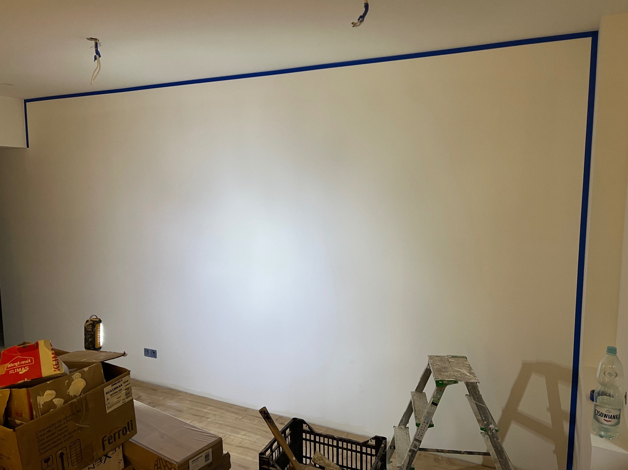 Przygotowana do malowania ściana w pomieszczeniu, zabezpieczona niebieską taśmą malarską wzdłuż sufitu i narożników, widoczne kartony z materiałami budowlanymi, drabina i oświetlenie warsztatowe
