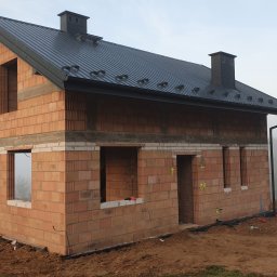 MANTER DANIEL KAZEK - Surowy stan nowego domu z czerwonej cegły ceramicznej, z czarnym dachem z blachy trapezowej, widoczne otwory okienne i drzwiowe, na zewnątrz mglista pogoda.