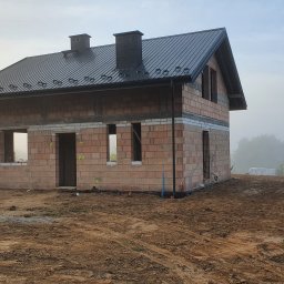MANTER DANIEL KAZEK - Surowy stan domu z czerwonej cegły ceramicznej z czarnym dachem blachodachówką w porannej mgle, widoczne niezamontowane okna i drzwi, teren wokół domu częściowo wyrównany.
