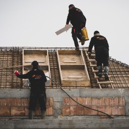 MANTER DANIEL KAZEK - Trzech robotników w czarnych kombinezonach z logo firmy budowlanej na budowie domu w trakcie wznoszenia ścian z czerwonej cegły i przygotowywania zbrojenia dachu, jeden z nich trzyma białą płytę...