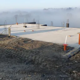 Zalana betonem płyta fundamentowa z wystającymi prętami zbrojeniowymi, widoczne deskowanie i poziomica, w tle zamglony krajobraz i fragment domu.