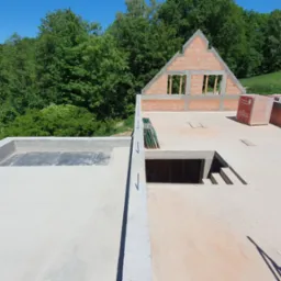 Wznoszenie ścian domu z pustaków ceramicznych na betonowej płycie fundamentowej z widocznym otworem na schody; w tle zielone drzewa i częściowo zabudowany teren.