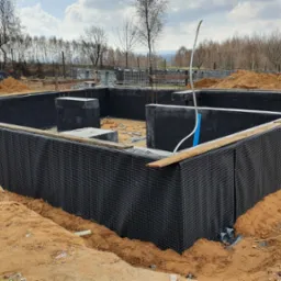 Przygotowany fundament z izolacją pionową z folii kubełkowej, zabezpieczony przed zasypaniem ziemią, widoczne zbrojenia i elementy konstrukcyjne wewnątrz, w tle drzewa i pagórkowaty teren.