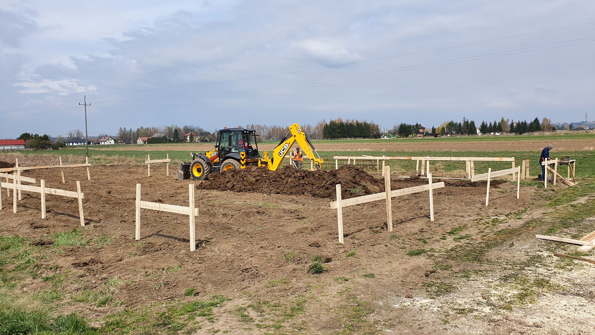Żółta koparko-ładowarka JCB pracuje przy wykopie pod fundamenty przyszłego domu na rozległym polu, widoczne drewniane szalunki i dwóch pracowników w oddali.