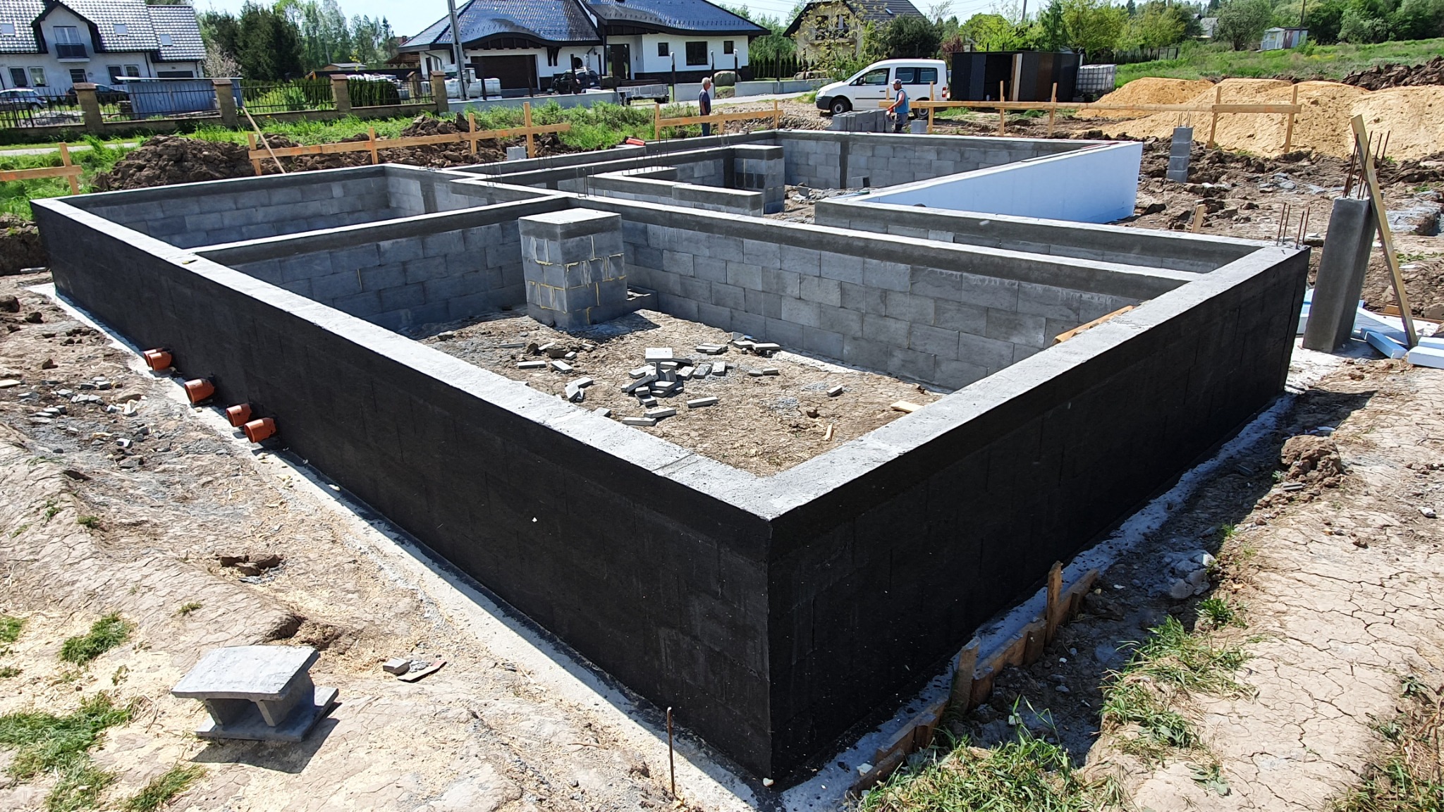 Wykopane fundamenty domu z izolacją przeciwwilgociową, widoczne bloczki betonowe, wystające pręty zbrojeniowe i pomarańczowe rury drenażowe.
