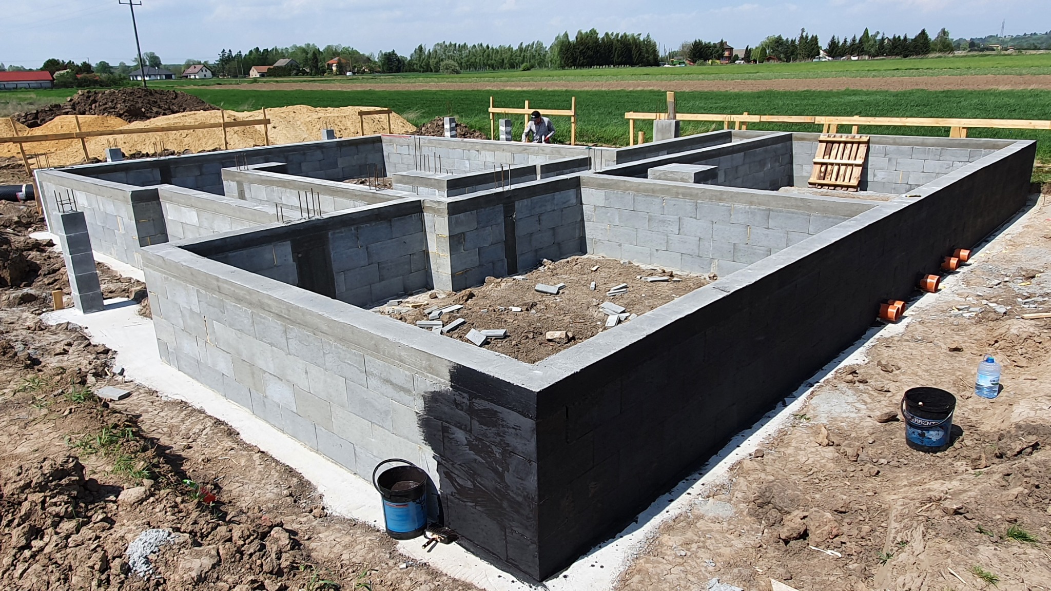 Fundamenty domu z bloczków betonowych, częściowo zabezpieczone czarną izolacją, widoczne wystające pręty zbrojeniowe, w tle krajobraz wiejski.