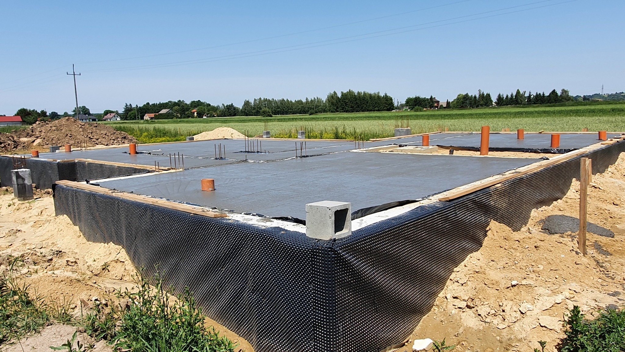 Zalany betonem fundament z hydroizolacją w postaci czarnej folii kubełkowej, widoczne wystające rury i pręty zbrojeniowe, otoczenie stanowi ziemia i trawa, w tle pole i zabudowania.