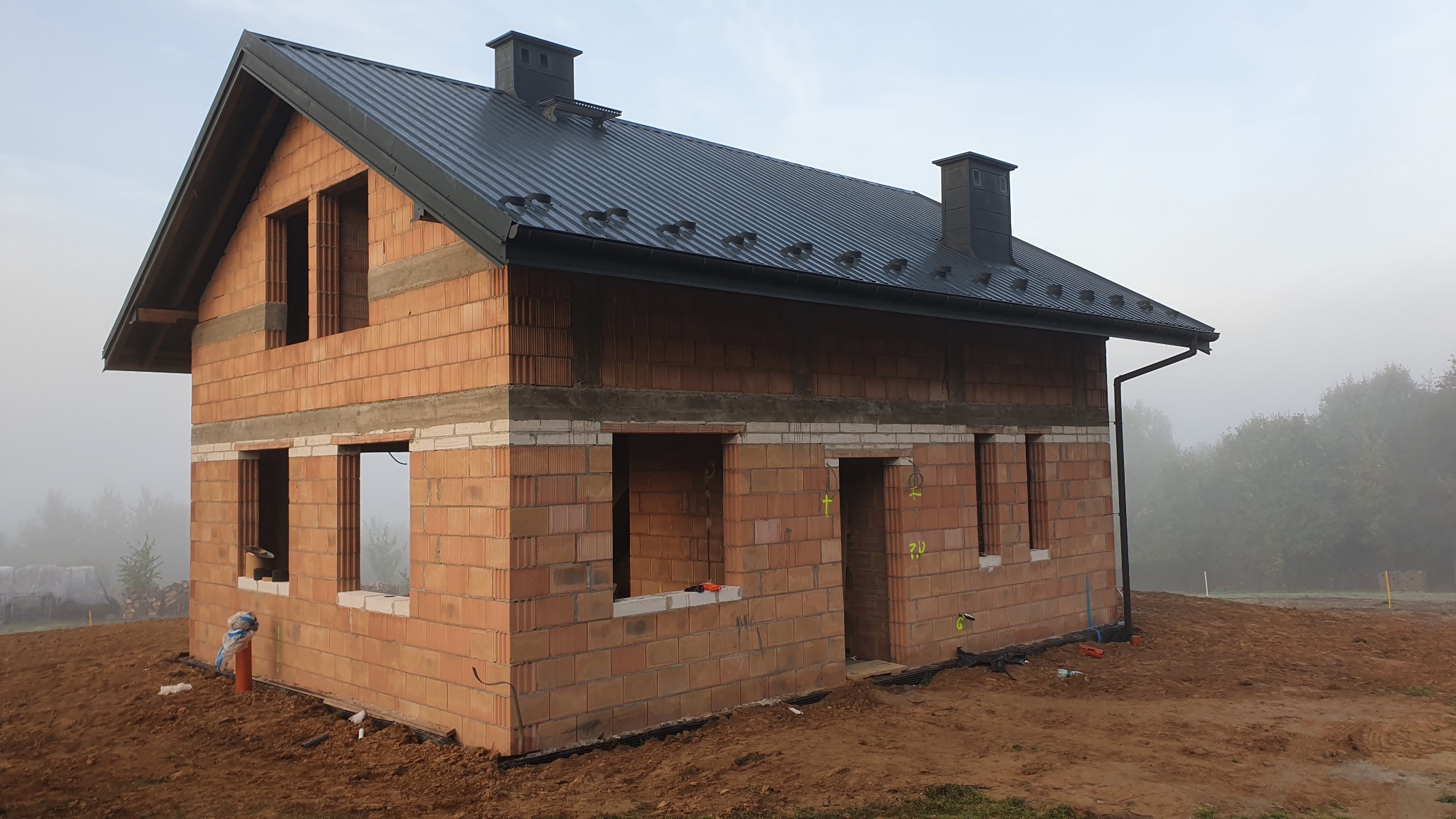 Surowy stan nowego domu z czerwonej cegły ceramicznej, z czarnym dachem z blachy trapezowej, widoczne otwory okienne i drzwiowe, na zewnątrz mglista pogoda.