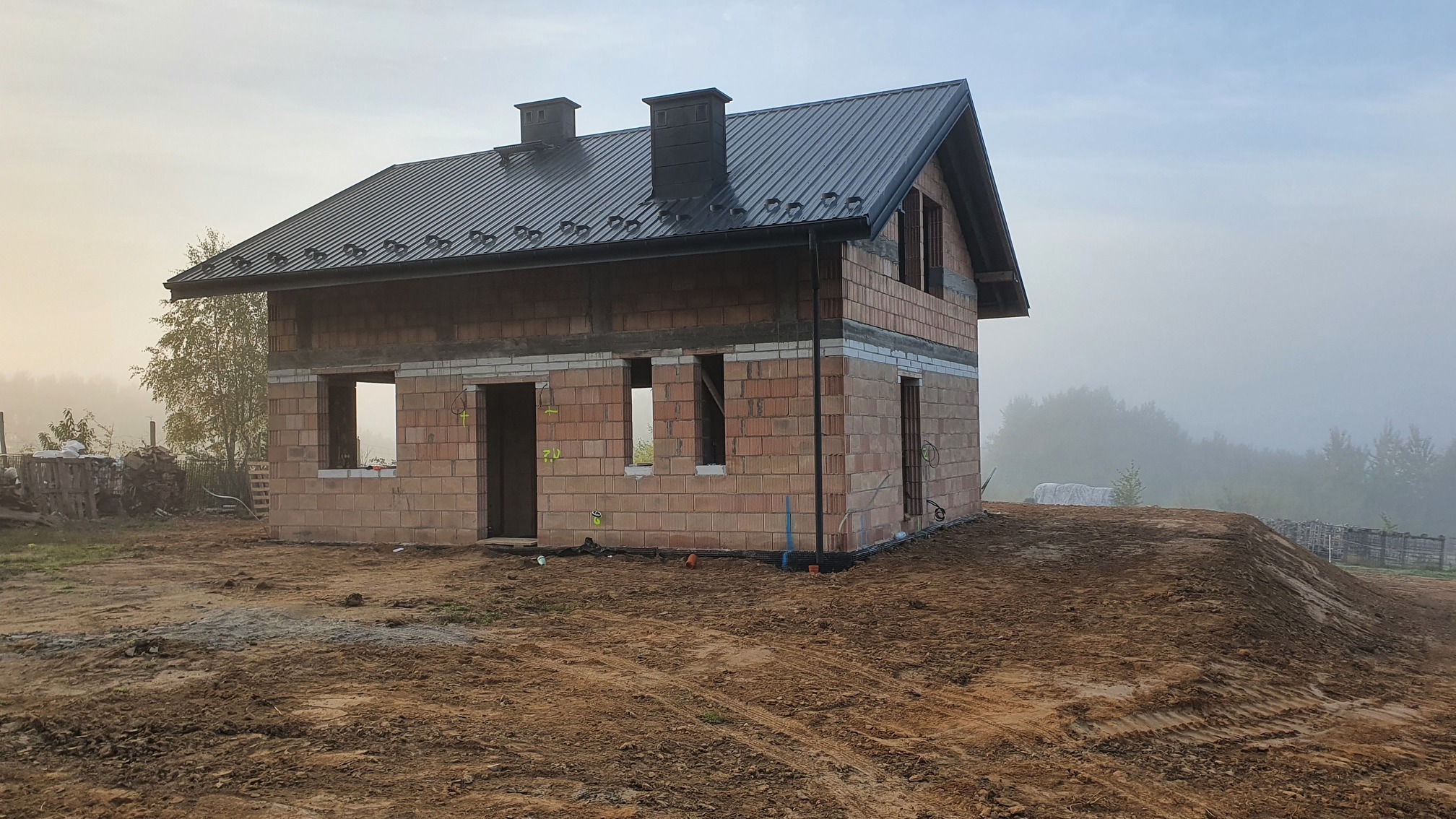 Surowy stan domu z czerwonej cegły ceramicznej z czarnym dachem blachodachówką w porannej mgle, widoczne niezamontowane okna i drzwi, teren wokół domu częściowo wyrównany.