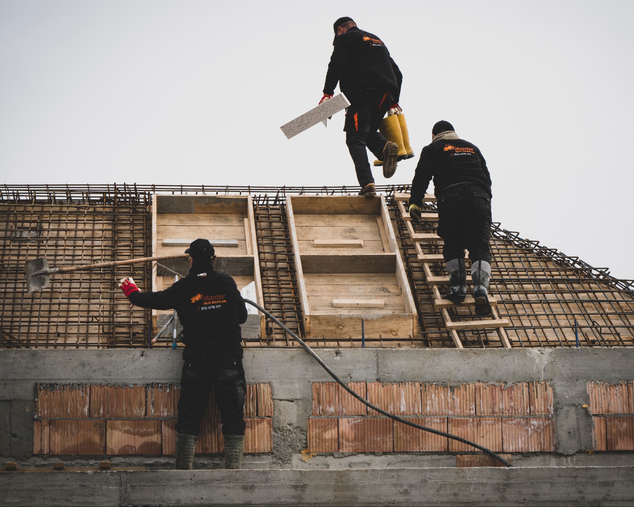 Trzech robotników w czarnych kombinezonach z logo firmy budowlanej na budowie domu w trakcie wznoszenia ścian z czerwonej cegły i przygotowywania zbrojenia dachu, jeden z nich trzyma białą płytę...