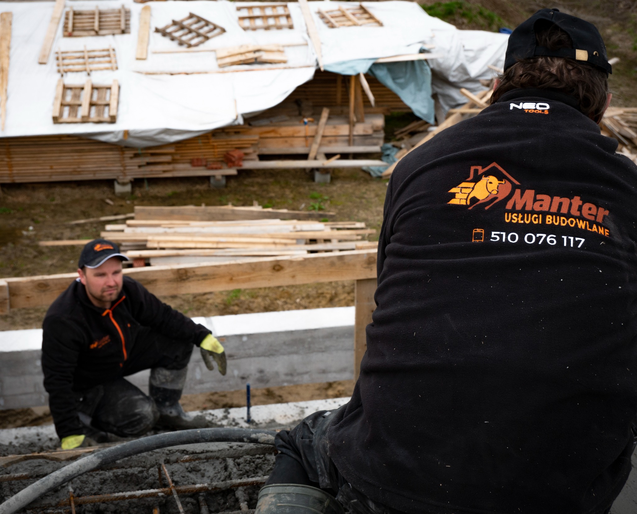 Dwóch robotników w czarnych bluzach z logo firmy Manter Usługi Budowlane pracuje przy wylewaniu fundamentów; widoczne zbrojenie, szalunek i przygotowane drewniane elementy konstrukcyjne w tle.