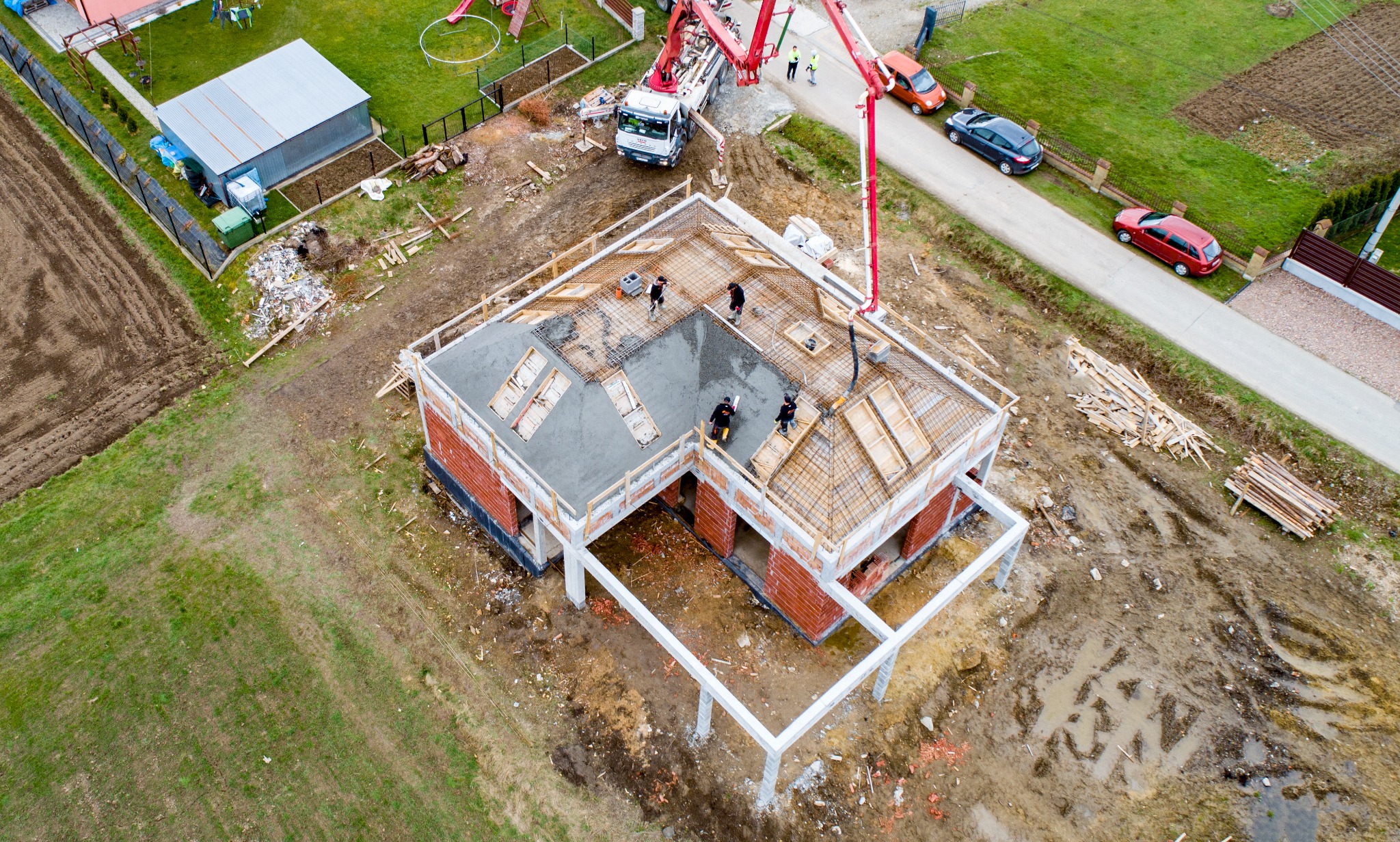 Widok z lotu ptaka na budowę domu z czerwonej cegły, z wylewanym betonem na stropie i pracownikami w czarnych ubraniach. Na zewnątrz stoi betonowóz z wysięgnikiem, a w tle widać domy i samochody.