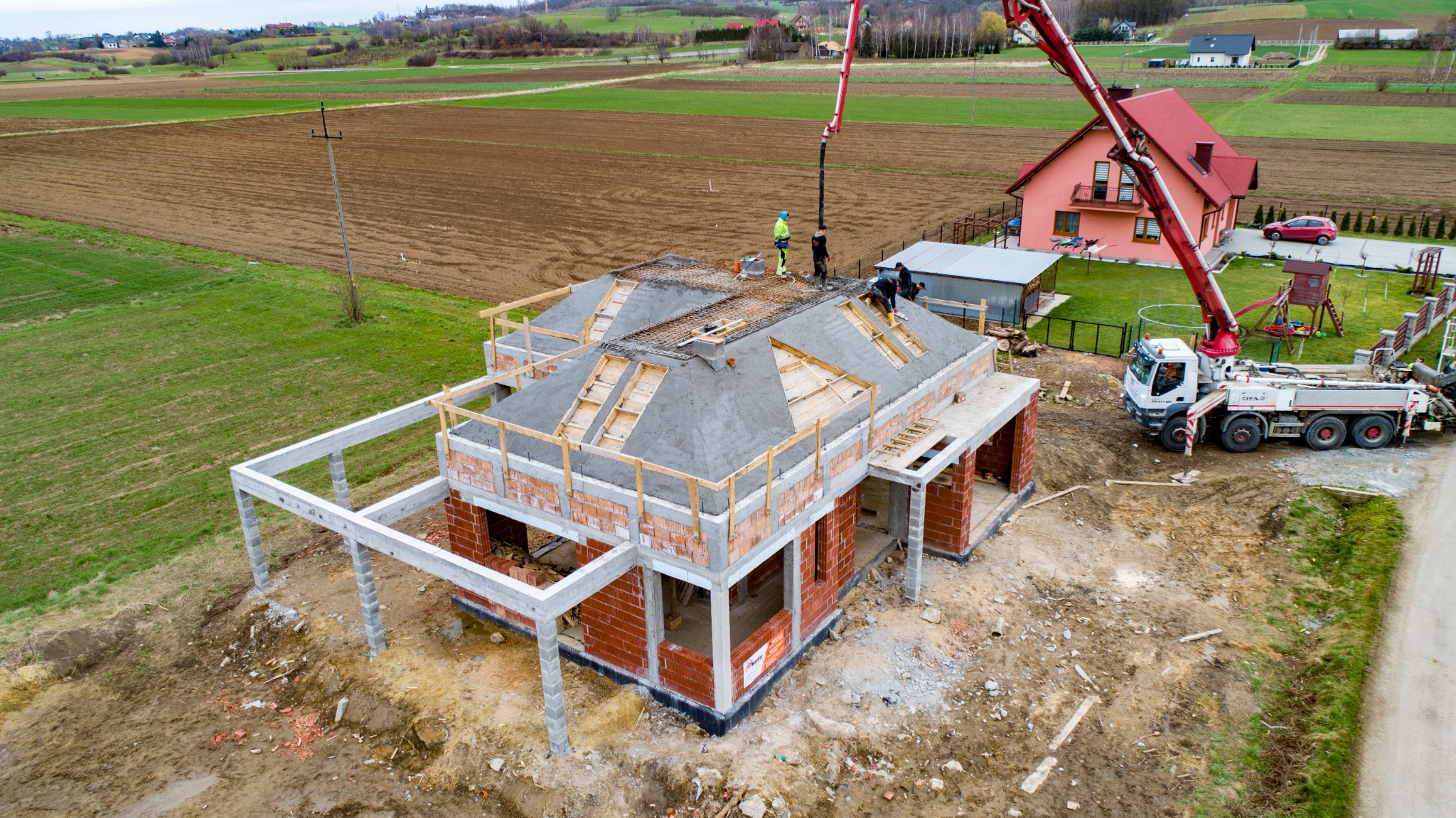 Wznoszenie konstrukcji domu jednorodzinnego z betonu komórkowego, widok z góry na budynek w trakcie wylewania betonowego stropu, z użyciem pompy do betonu. W tle pola uprawne i zabudowa jednorodzinna.