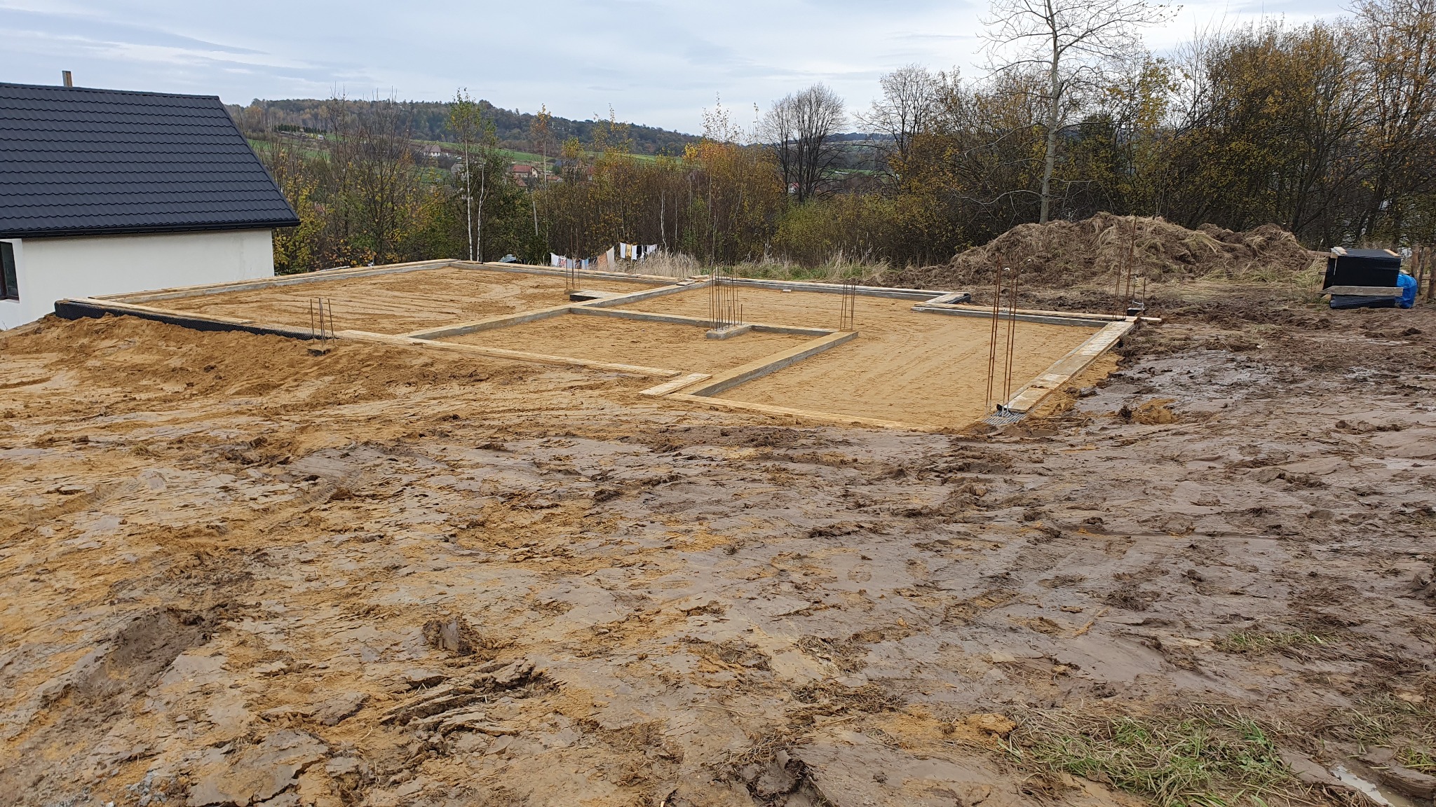 Przygotowany teren pod budowę domu z widocznymi fundamentami w szalunkach i wystającymi prętami zbrojeniowymi, otoczony błotnistym terenem i zalesionym wzgórzem w tle.