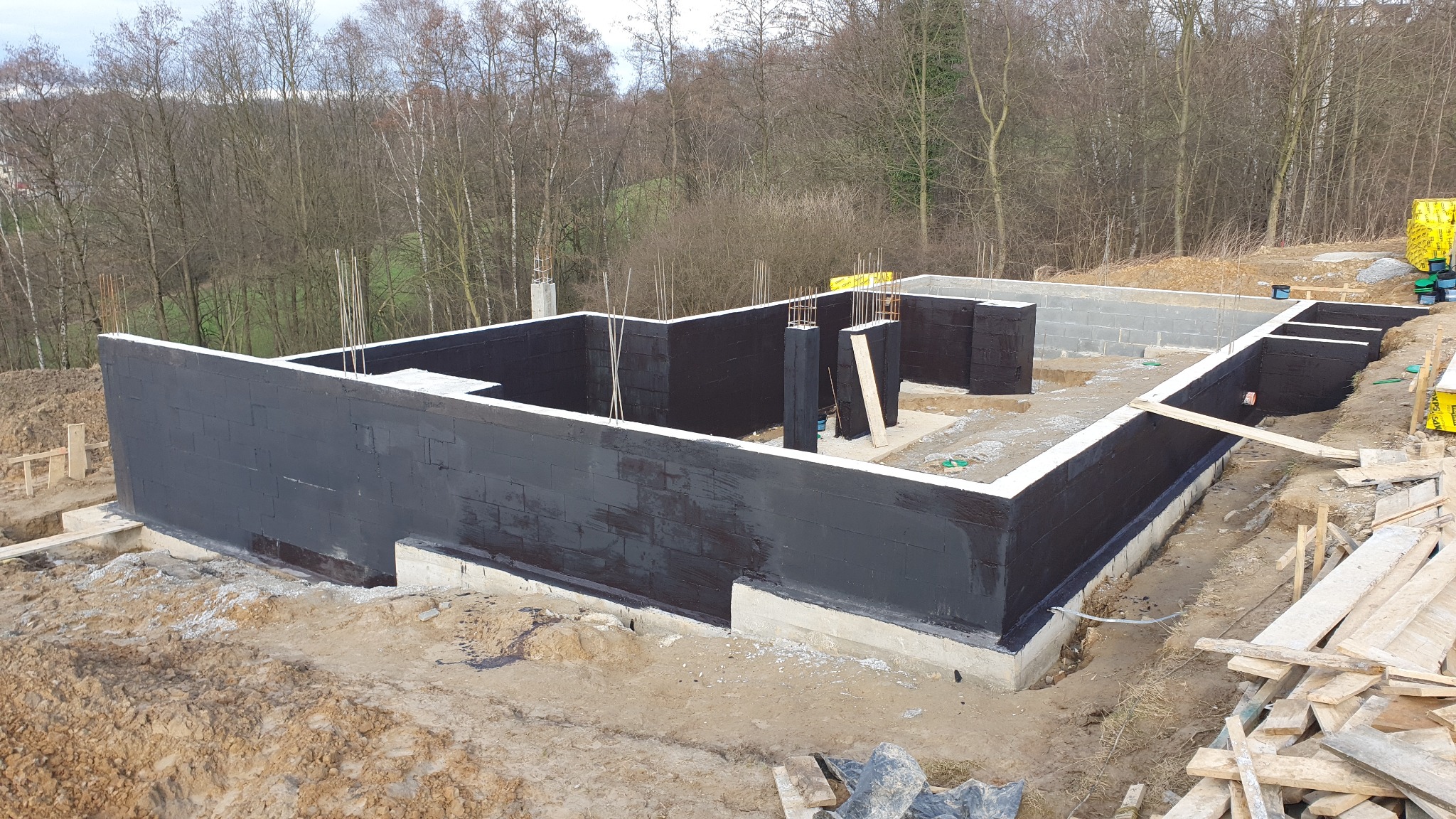 Fundamenty domu z bloczków betonowych, zabezpieczone czarną izolacją bitumiczną, widoczne pręty zbrojeniowe i teren budowy z drewnianymi elementami szalunku.