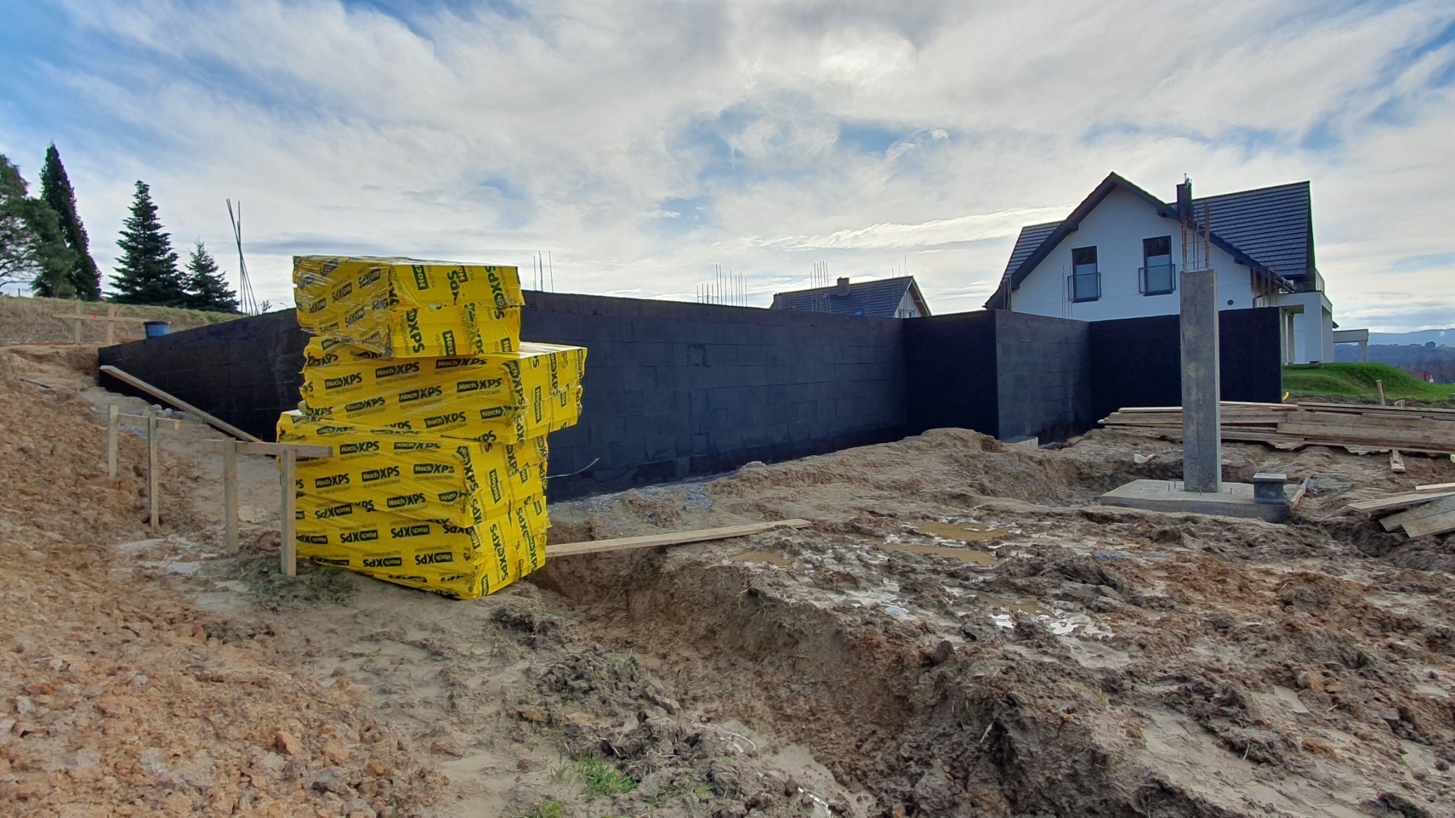 Przygotowany teren pod budowę domu w Mokrzysku z widocznymi fundamentami zabezpieczonymi czarną izolacją oraz stosem żółtych płyt izolacyjnych XPS w opakowaniu, w tle widoczny budynek mieszkalny.