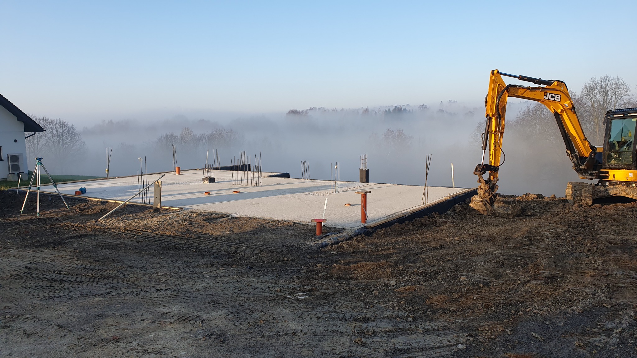 Wylana płyta fundamentowa z wystającymi prętami zbrojeniowymi i pionowymi rurami instalacyjnymi, widoczny żółty traktor JCB i niwelator laserowy na trójnogu, w tle mglista panorama.