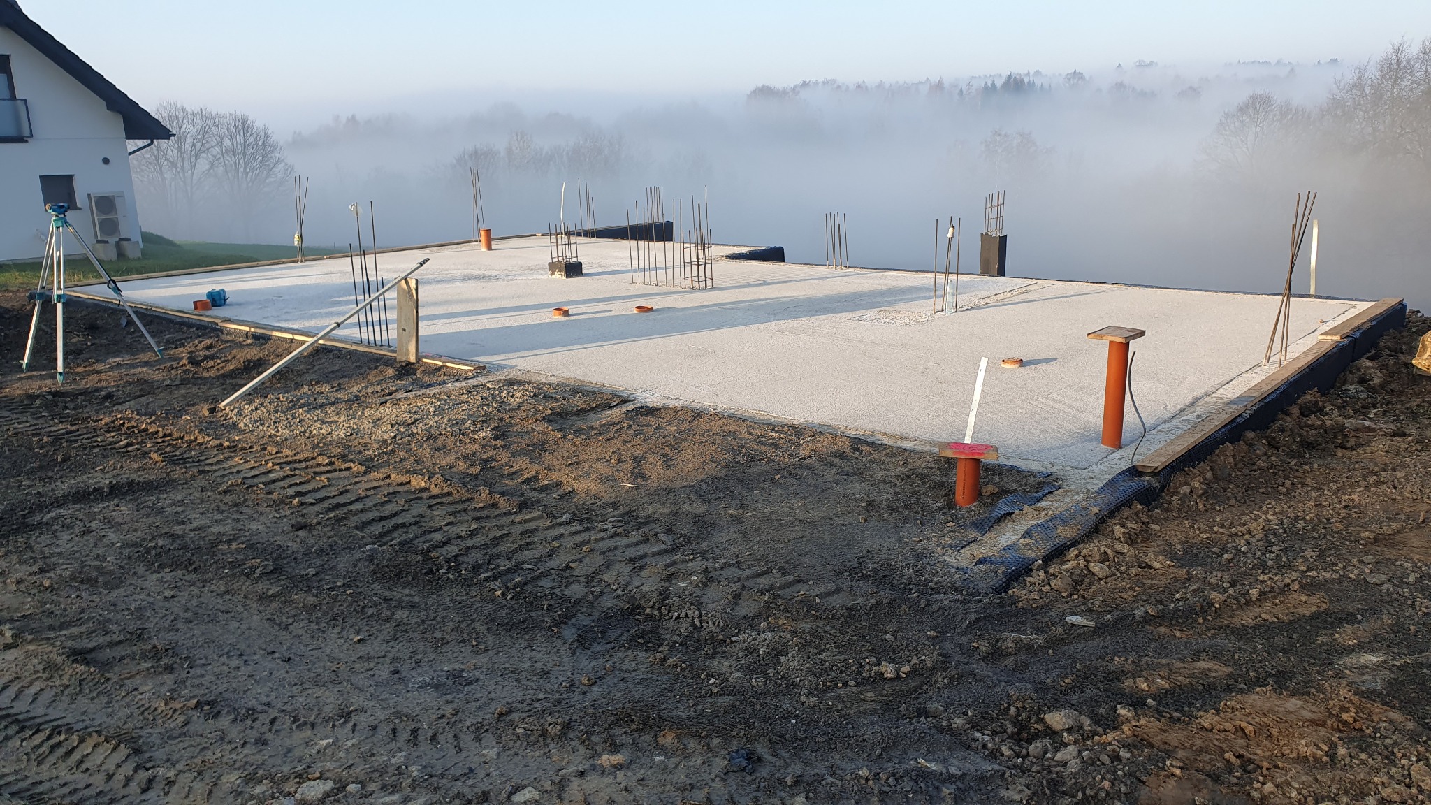 Zalana betonem płyta fundamentowa z wystającymi prętami zbrojeniowymi, widoczne deskowanie i poziomica, w tle zamglony krajobraz i fragment domu.