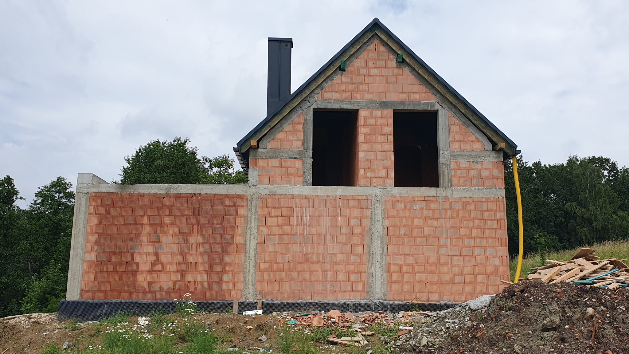 Surowy stan budynku w trakcie budowy z czerwonej cegły ceramicznej, widoczne otwory okienne i komin, na pierwszym planie składowane drewno budowlane, otoczenie naturalne.