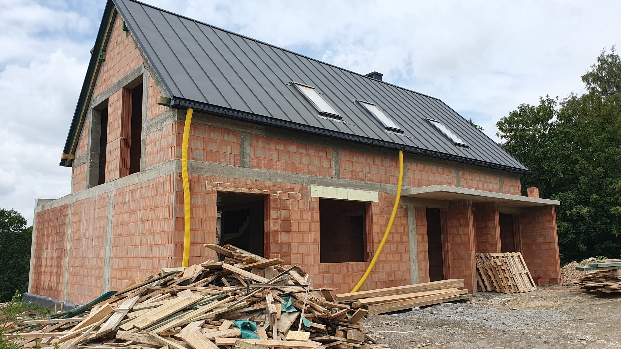 Surowy budynek z czerwonej cegły z ciemnoszarym dachem i trzema oknami dachowymi, widoczne żółte rury spustowe, sterta drewna i palet przed budynkiem, budowa w toku.
