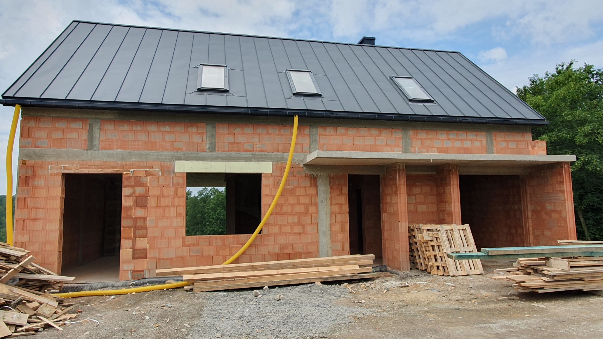 Surowy stan domu z czerwonej cegły z szarym dachem i trzema oknami dachowymi, widoczne drewniane palety i rura spustowa na zewnątrz.