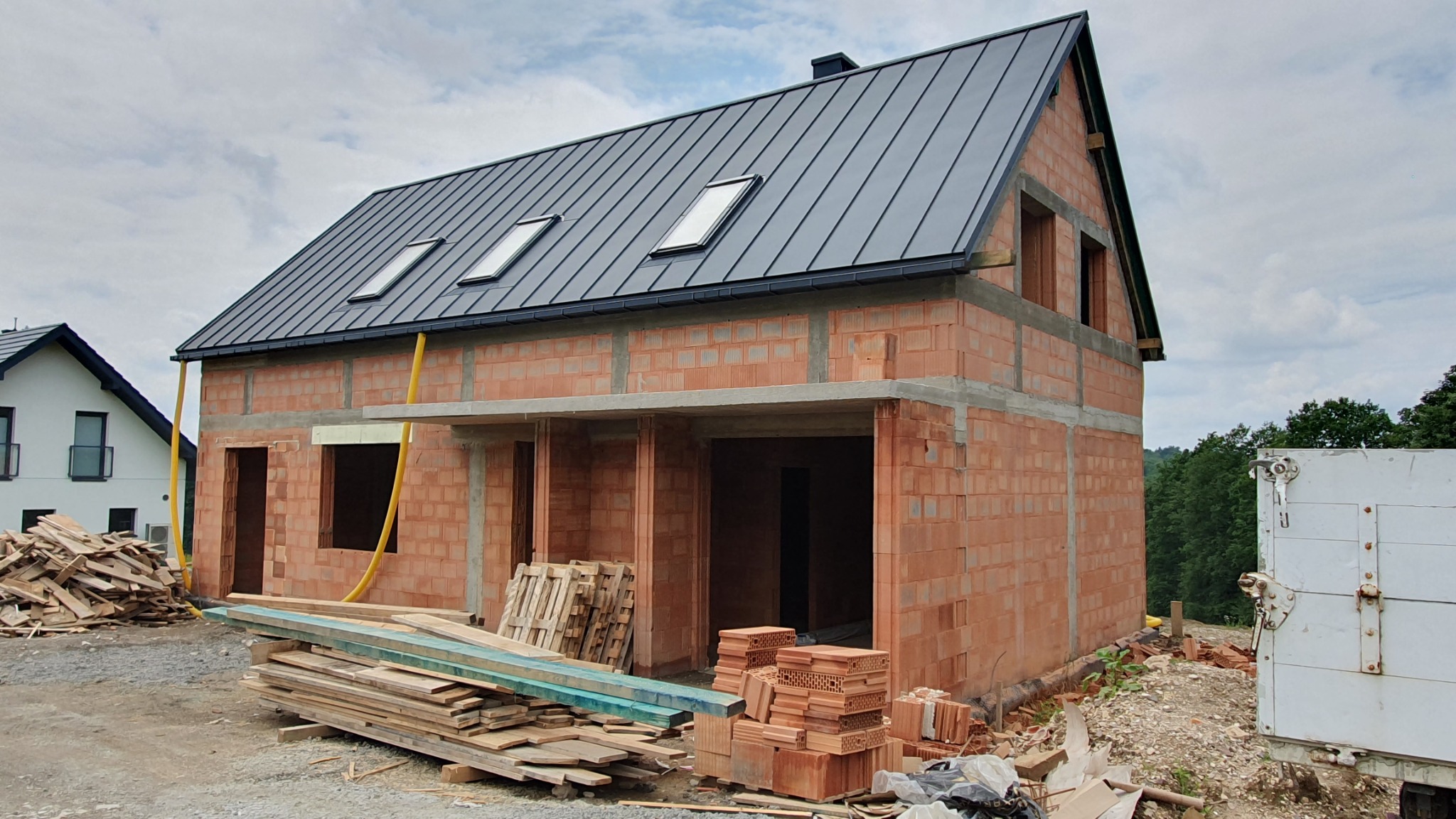 Surowy, dwukondygnacyjny dom murowany z czerwonej cegły, z ciemnoszarą, metalową dachówką i trzema oknami dachowymi, w trakcie budowy, z widocznymi elementami konstrukcyjnymi, drewnem i pustakami...