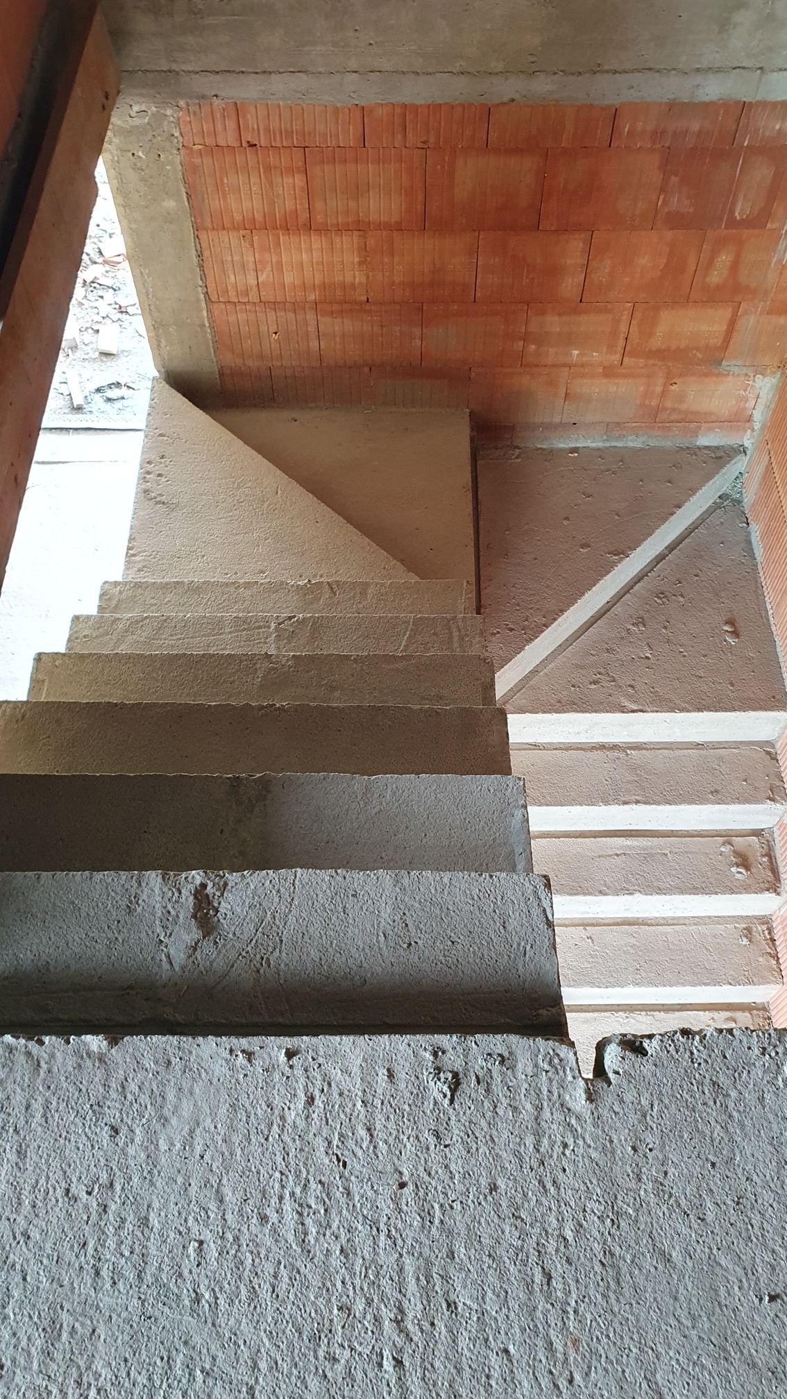Surowe, betonowe schody w trakcie budowy, z widocznymi fragmentami ceglanej ściany i surowego betonu, uchwycone z perspektywy góry.